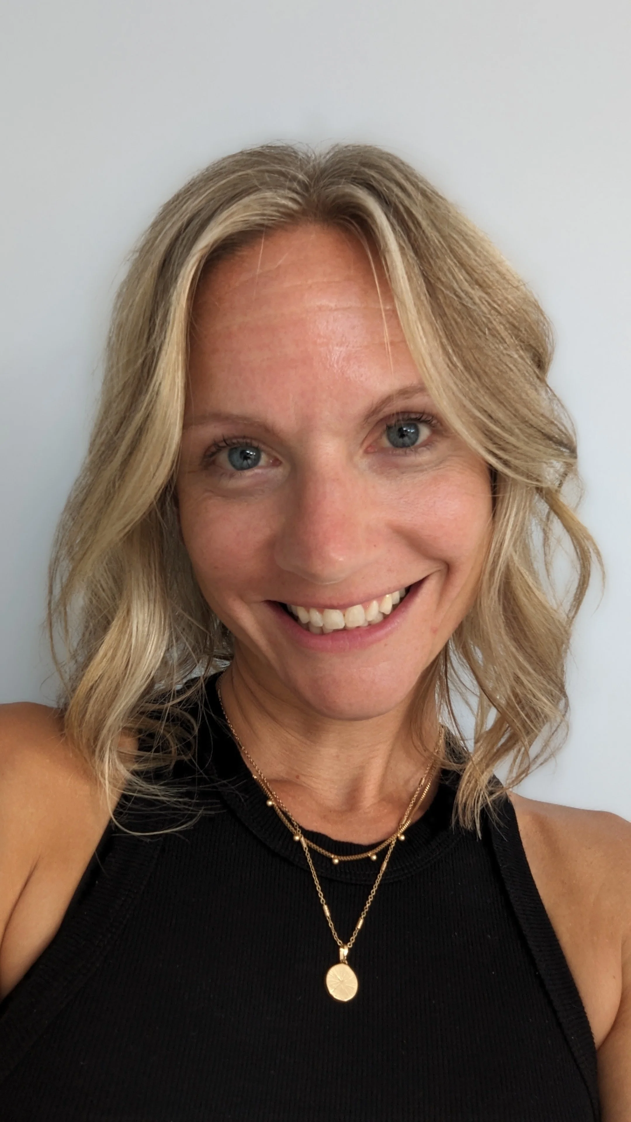 A woman with blonde hair in loose waves, smiling at the camera against a plain light background, wearing a black sleeveless top and layered gold necklaces.