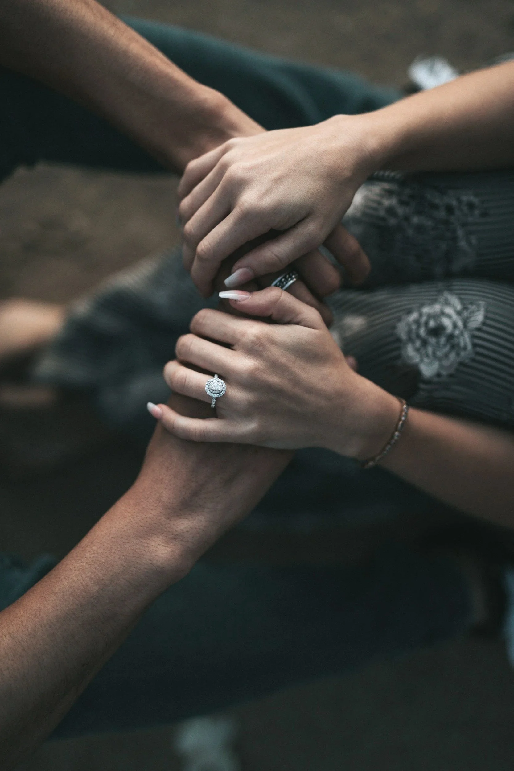 Two people holding hands, one with darker skin and the other with lighter skin, wearing rings, with a dark background.
