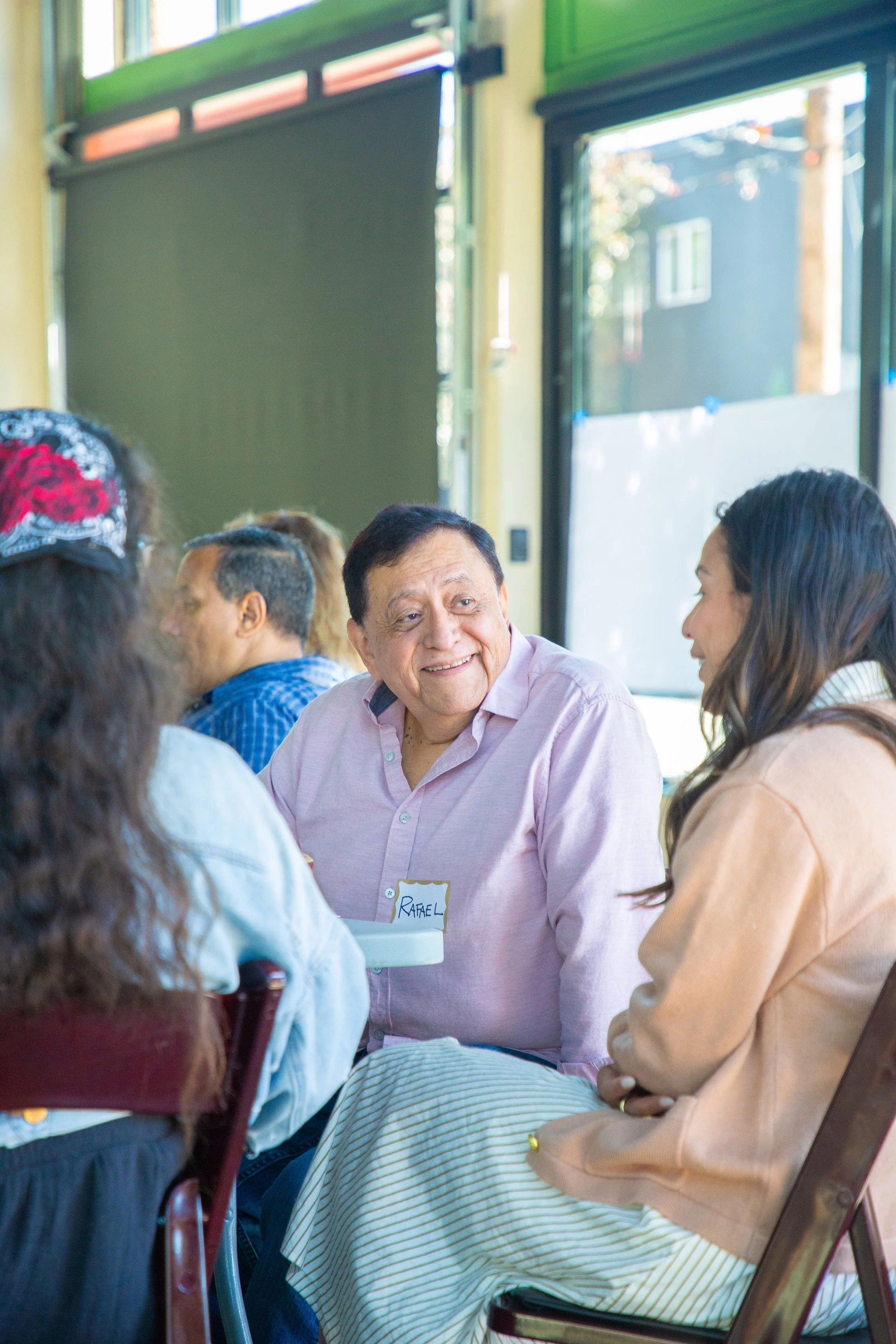 A group of people gathered around a table, engaging in a conversation, with a man wearing a pink shirt smiling, and a woman in a beige jacket listening attentively. The scene is indoors with large windows allowing natural light in.
