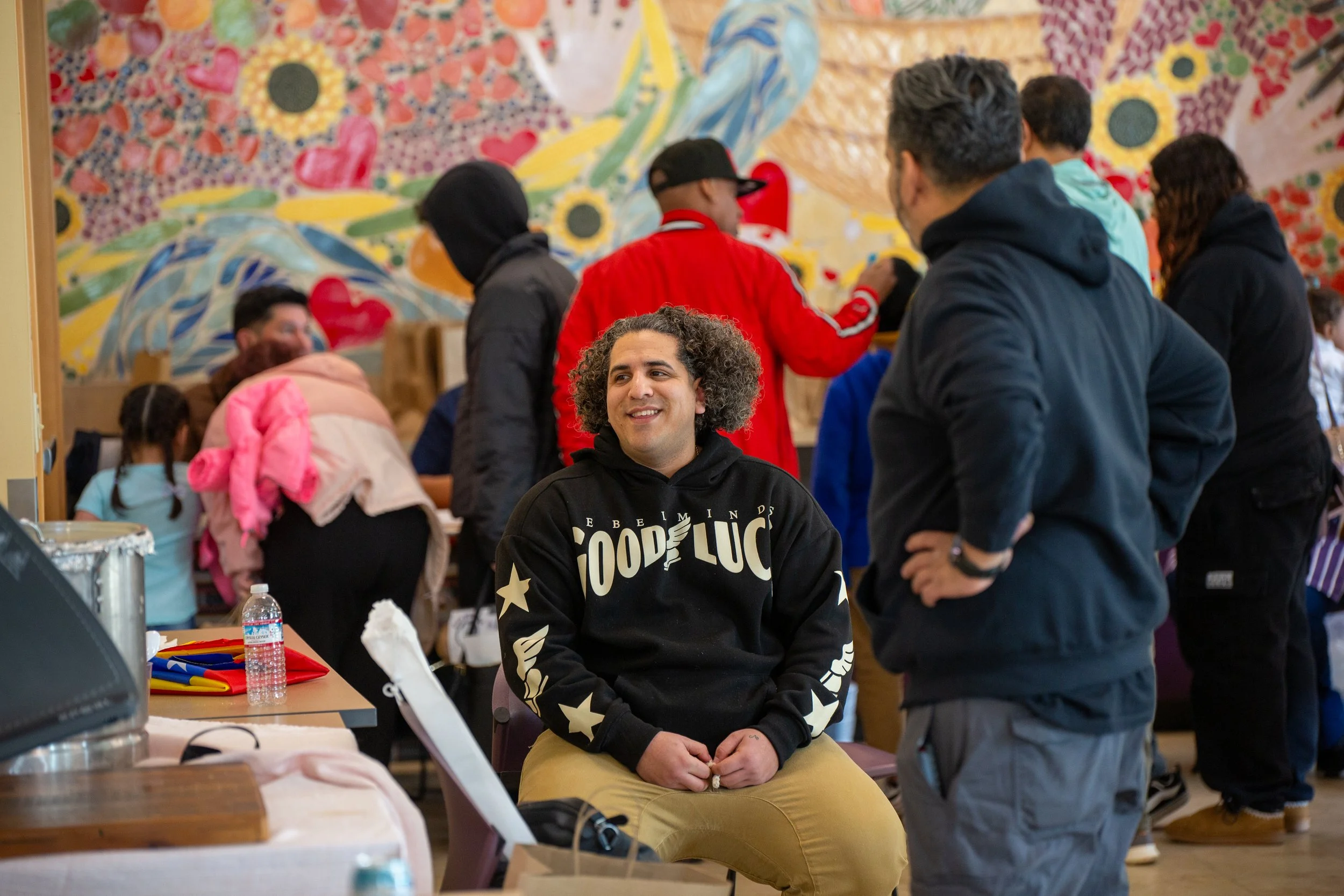 A young man with curly hair wearing a black hoodie with white lettering, smiling and sitting in a busy indoor space with people standing and talking around him, and colorful artwork on the wall in the background.