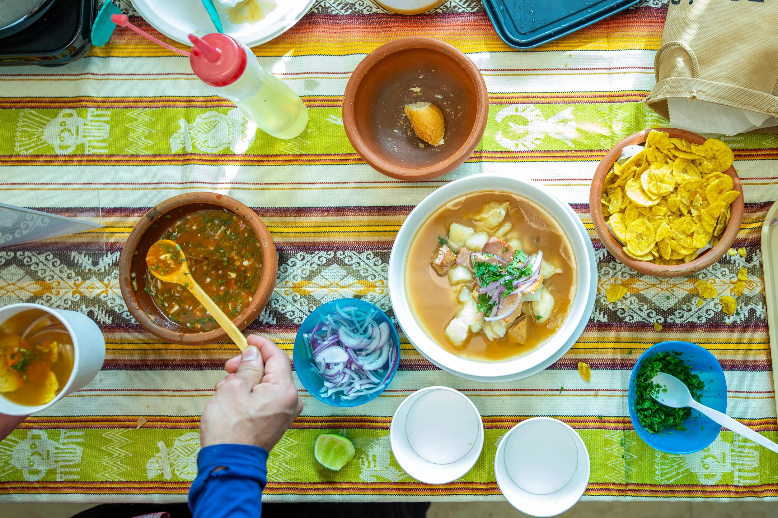 Top-down view of a colorful table with various Mexican dishes including bowls of soup, chopped onions, cilantro, a bowl of chips, and condiments, with a person's hand holding a spoonful of soup.