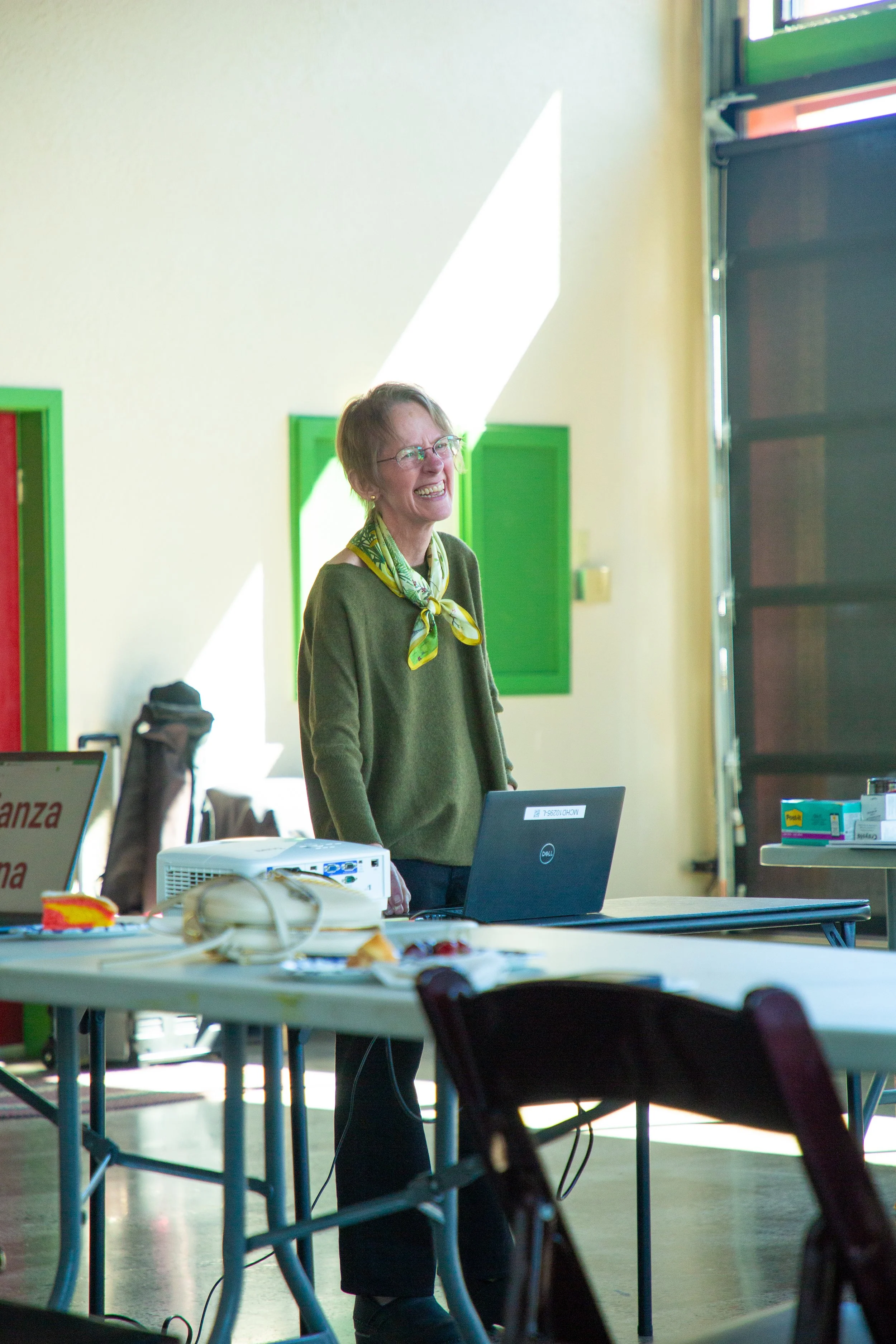 A woman with short light brown hair, glasses, and a yellow scarf is standing in a room with green framed windows and a yellow wall. She is smiling and appears to be speaking or presenting. There are tables with laptops, papers, and office supplies, including a projector and a box of tissues, in the room.
