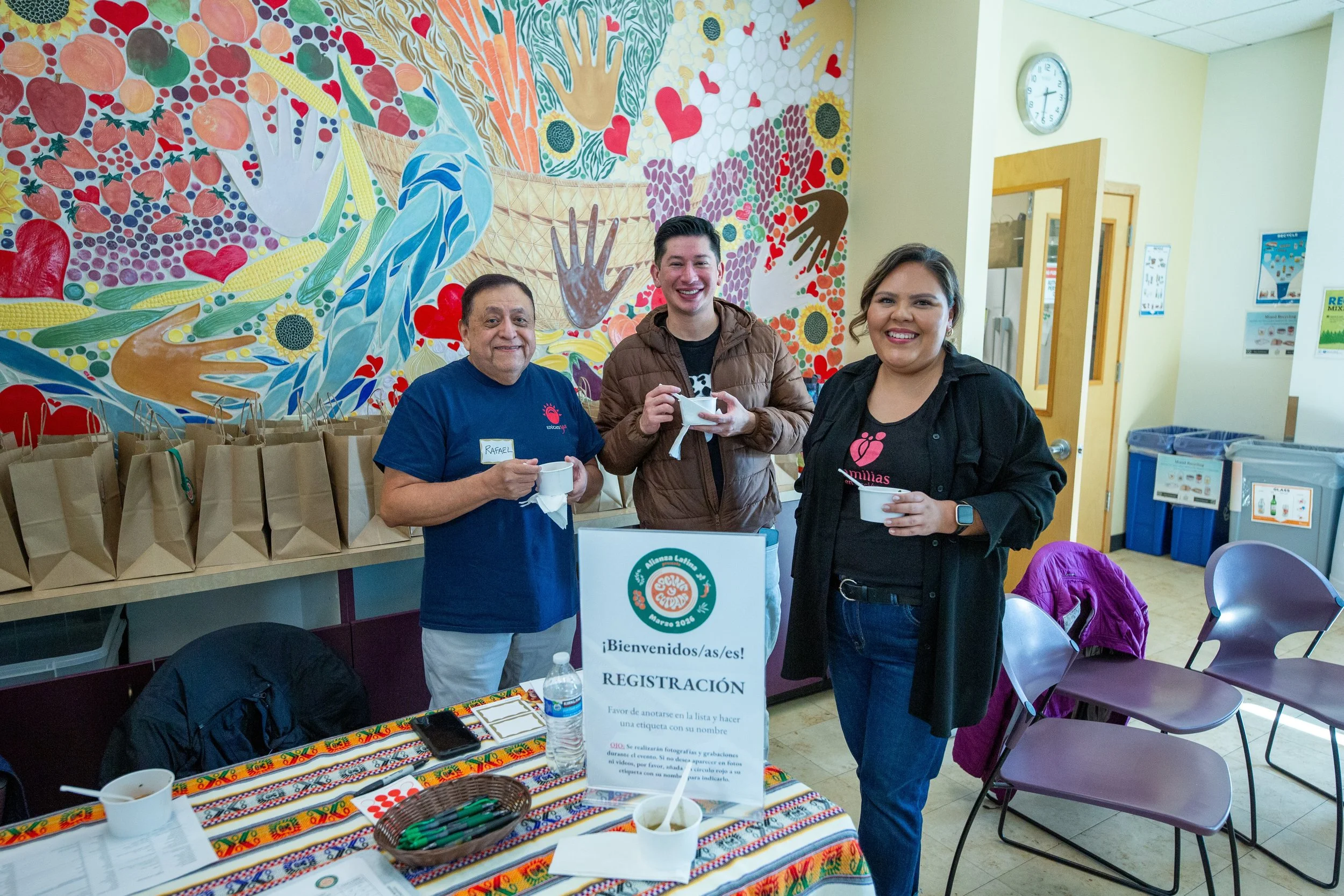 Three people standing indoors, smiling, near a registration table with a colorful tablecloth, cups, and papers. Behind them, a vibrant mural with various hearts, flowers, hands, and abstract designs covers the wall. Paper gift bags are lined up on a counter, and a sign indicates registration in Spanish.