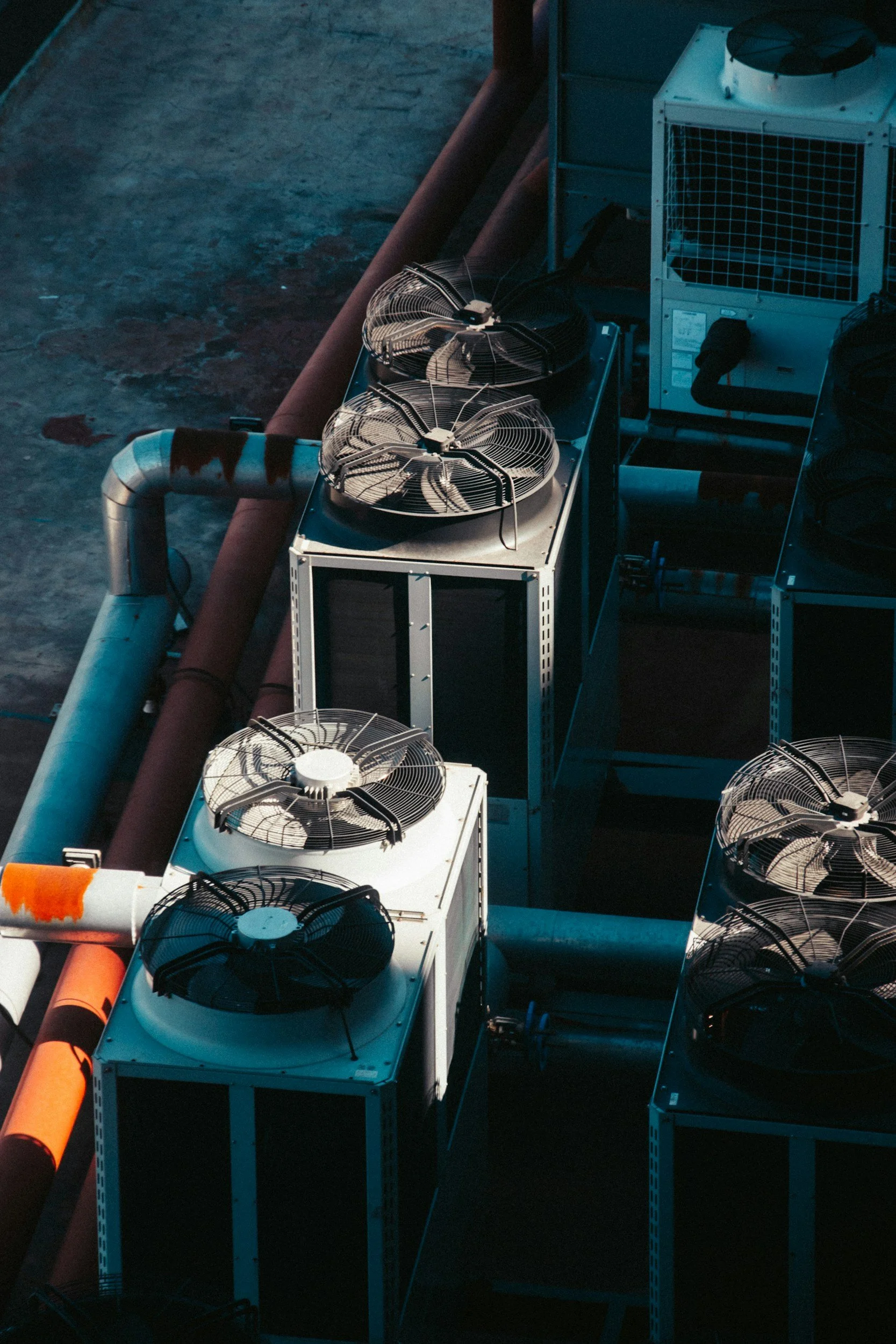 View of a rooftop HVAC system with multiple large fans and duct pipes, some orange and blue, against a dark, possibly evening sky.