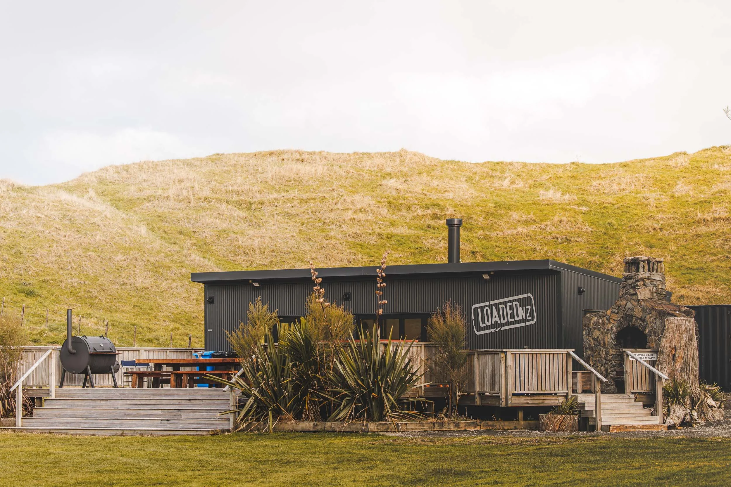 A modern black building with a sign that reads 'LOADENZ' is situated in front of a grassy hillside under a cloudy sky. The yard includes outdoor furniture, a grill, and plants.