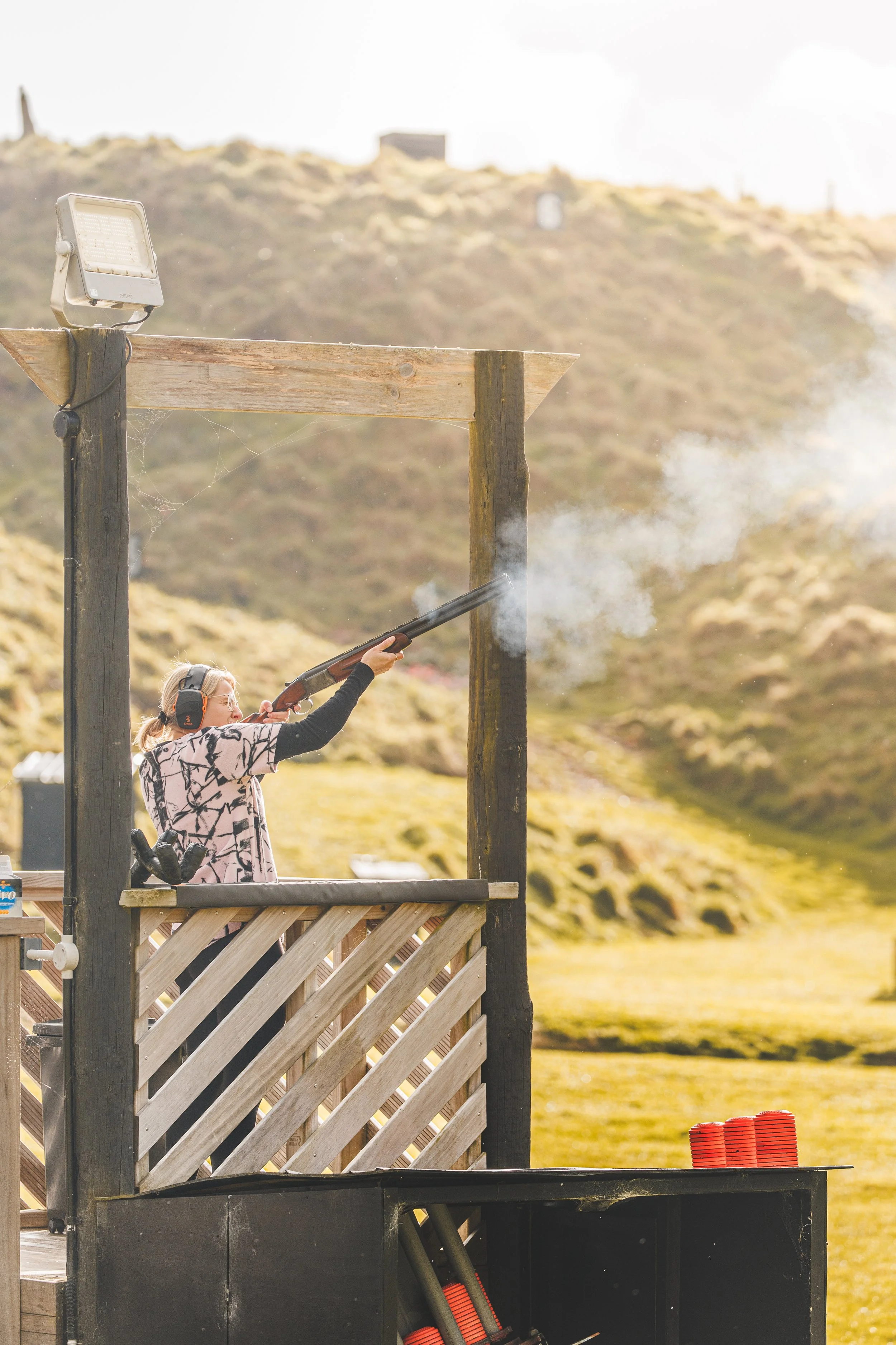 A woman wearing headphones and glasses is aiming a shotgun during target practice at an outdoor shooting range. She is standing on a wooden platform with a railing, and there are red clay targets stacked to her right. The background features a hilly landscape with sparse vegetation and a clear sky.