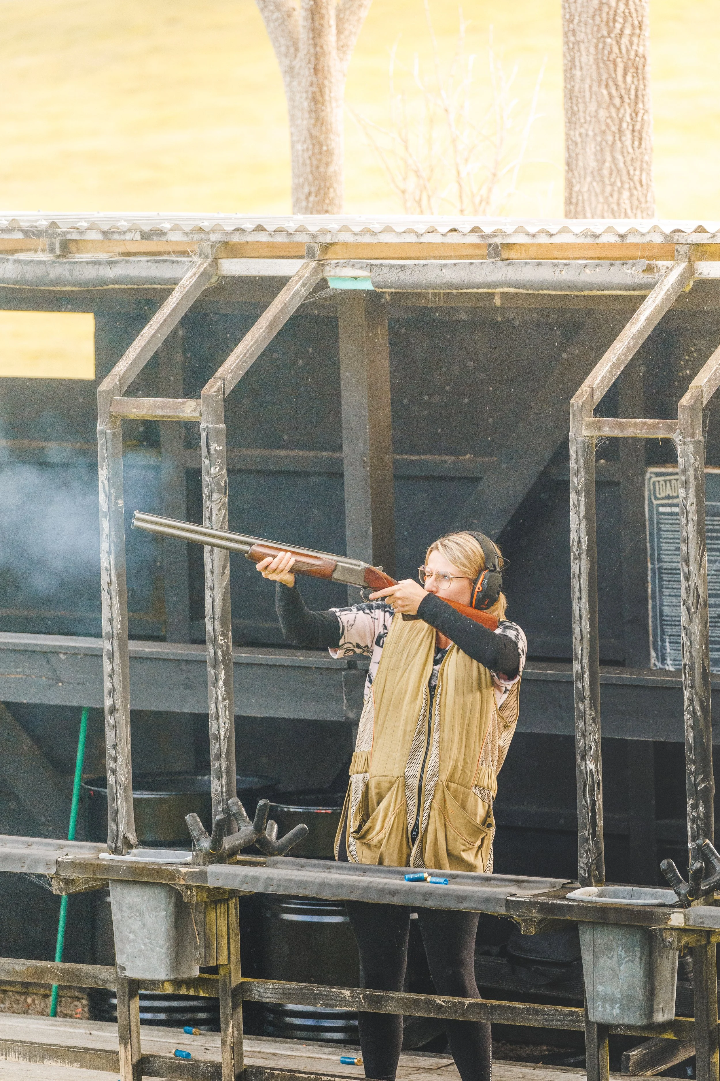 A woman at a shooting range holding a rifle and aiming, wearing protective earmuffs, glasses, a tan vest, black leggings, and a long-sleeve shirt with patterns, standing behind a metal shooting booth with an outdoor background of trees and a yellow sky.