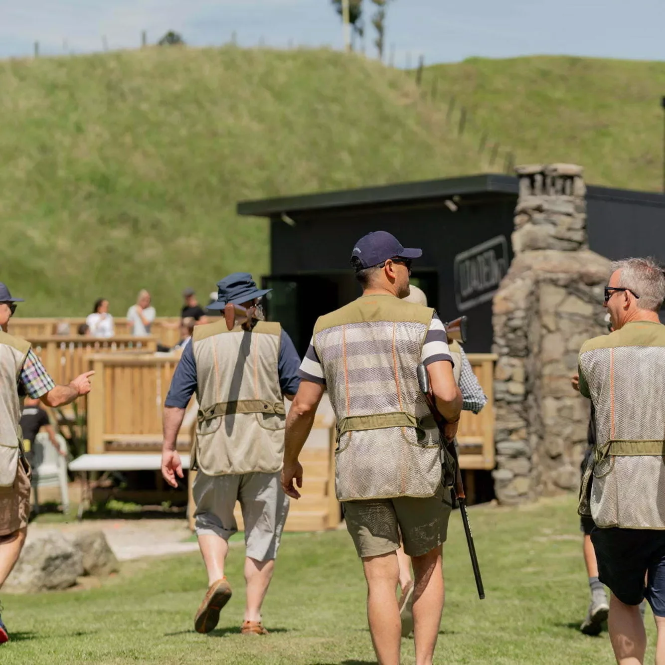 People participating in clay shooting, wearing vests and hats, holding shotguns, on a grassy outdoor range with a green hill in the background.
