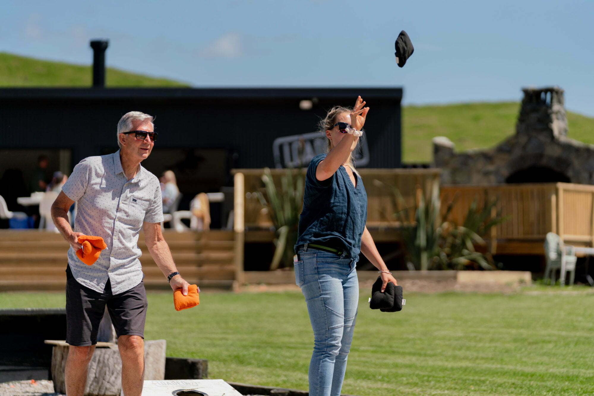 An older man and a younger woman at an outdoor event, throwing black and orange bean bags towards a cornhole board. The woman is wearing sunglasses and a blue shirt with jeans, while the man is in a white patterned shirt and dark shorts.