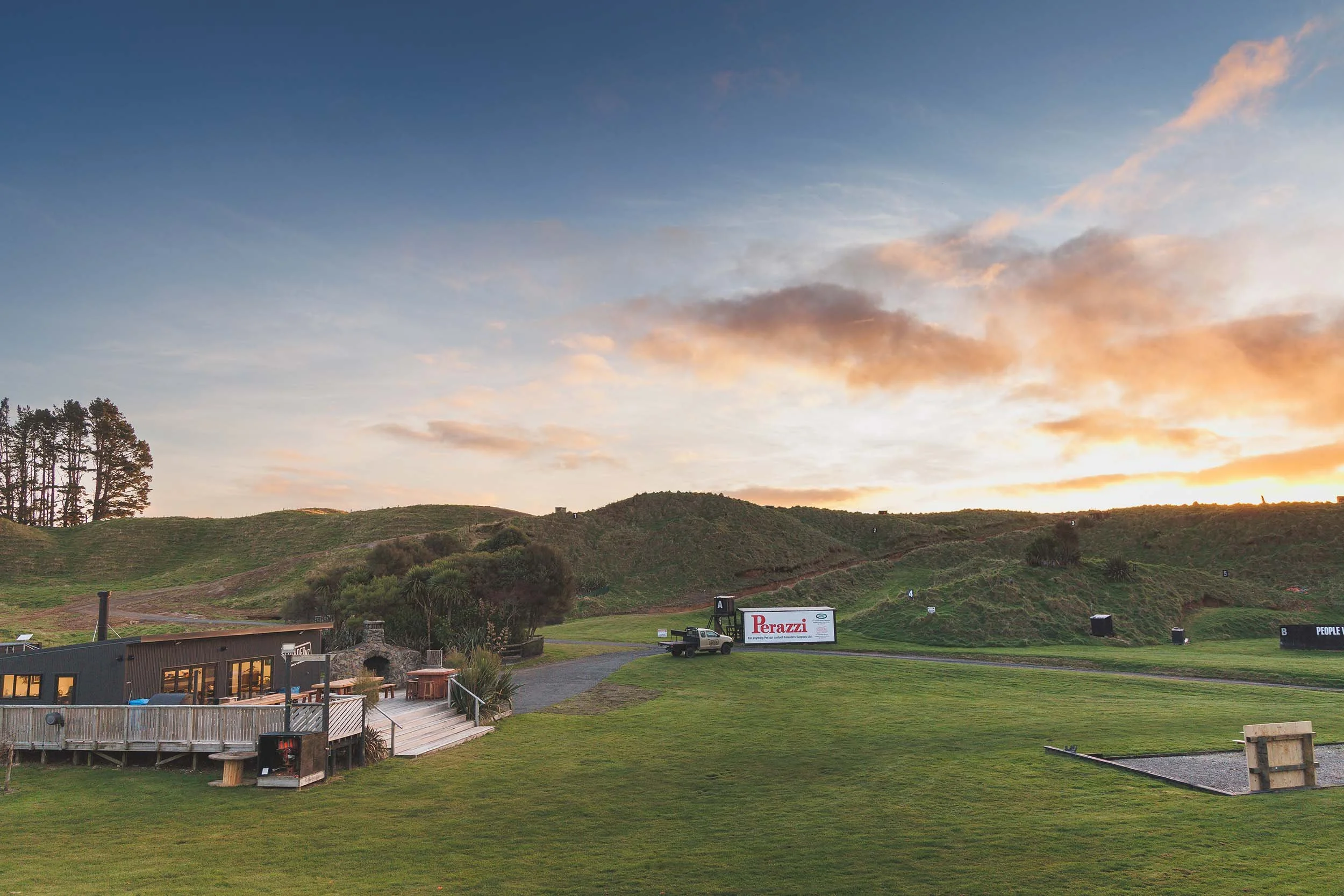 Scenic view of the Loaded NZ rolling landscape with trees, grass, a building, and a billboard, under a colourful sunset sky with scattered clouds.