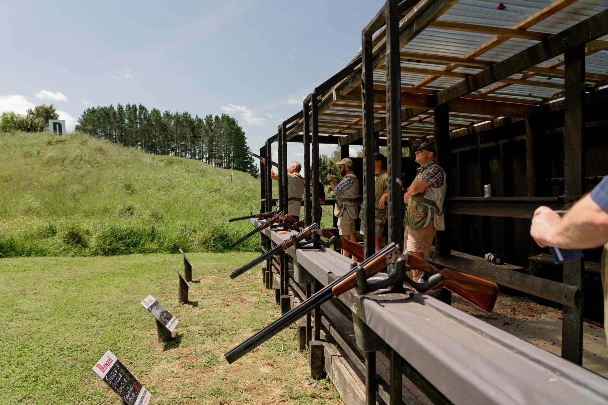 People at an outdoor shooting range with rifles aimed at targets down the hill, under a wooden and metal shelter.