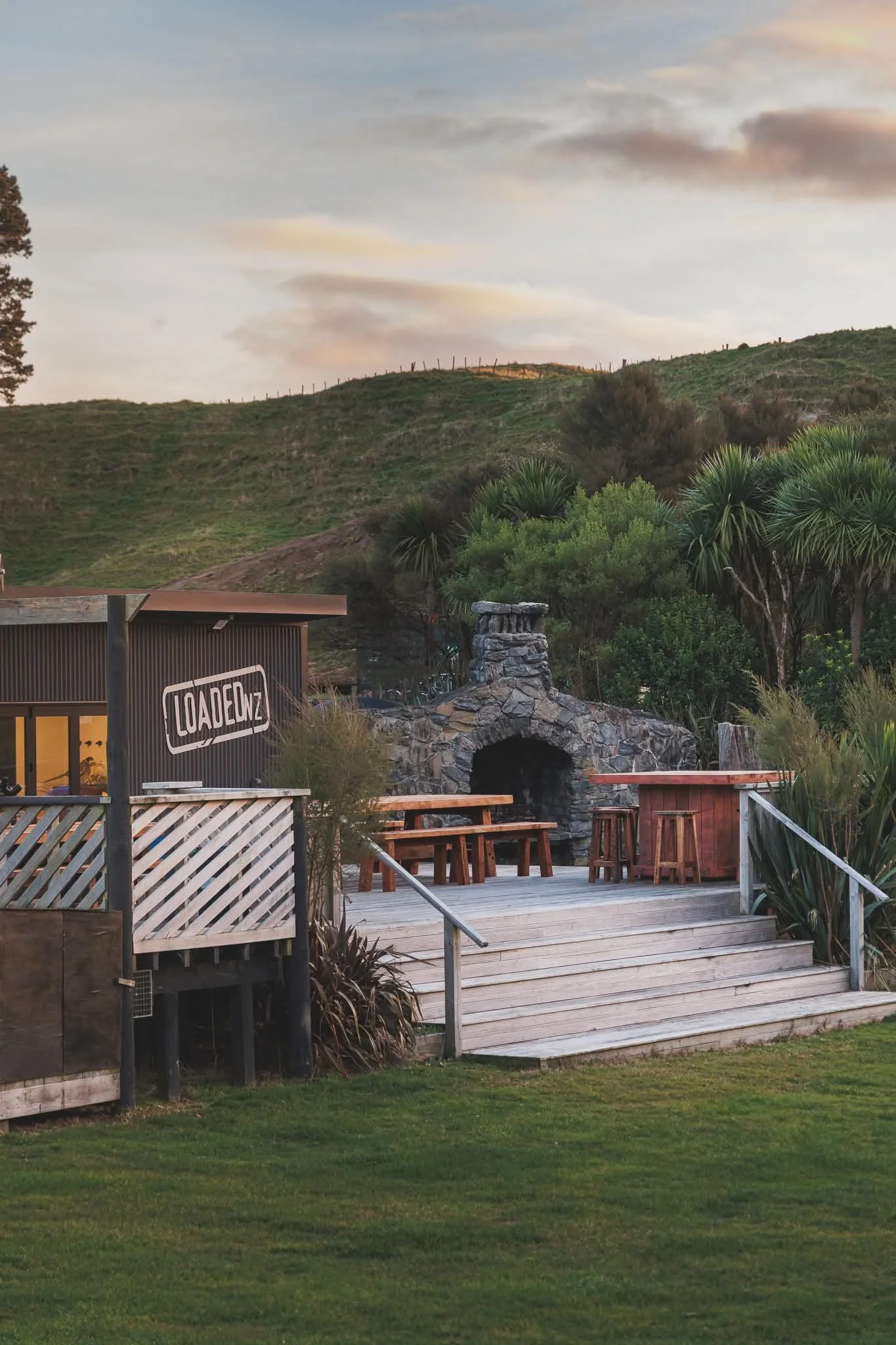 An outdoor patio area with wooden stairs and a deck, surrounded by green grass and plants, with a hillside and cloudy sky in the background.