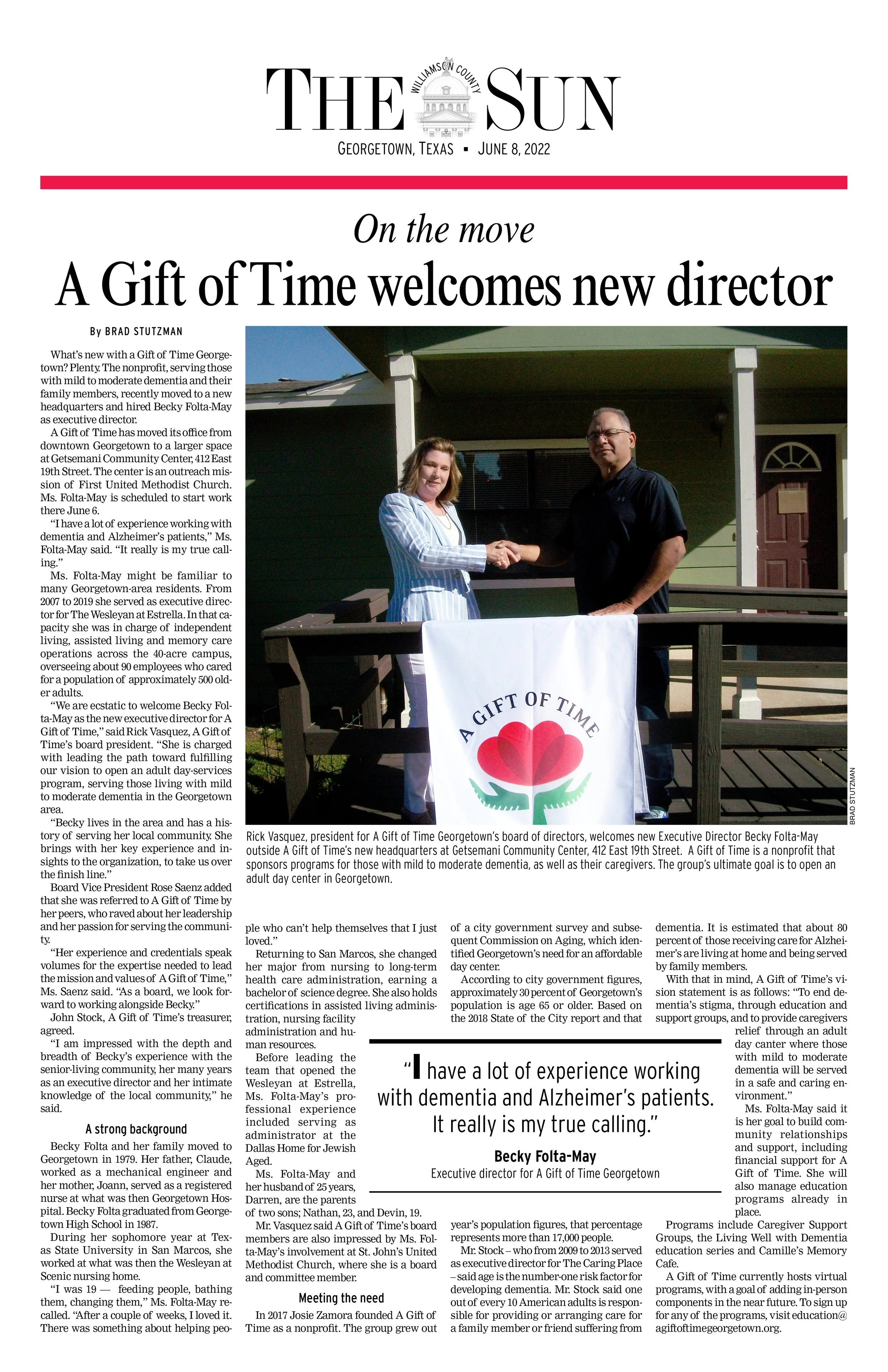 Photo of a woman and a man shaking hands outside during a ceremony in front of a small green building with a porch. The woman is wearing a light-colored striped suit and the man is in dark clothing. There is a banner with a red flower logo and the text "A Gift of Time" on the table.