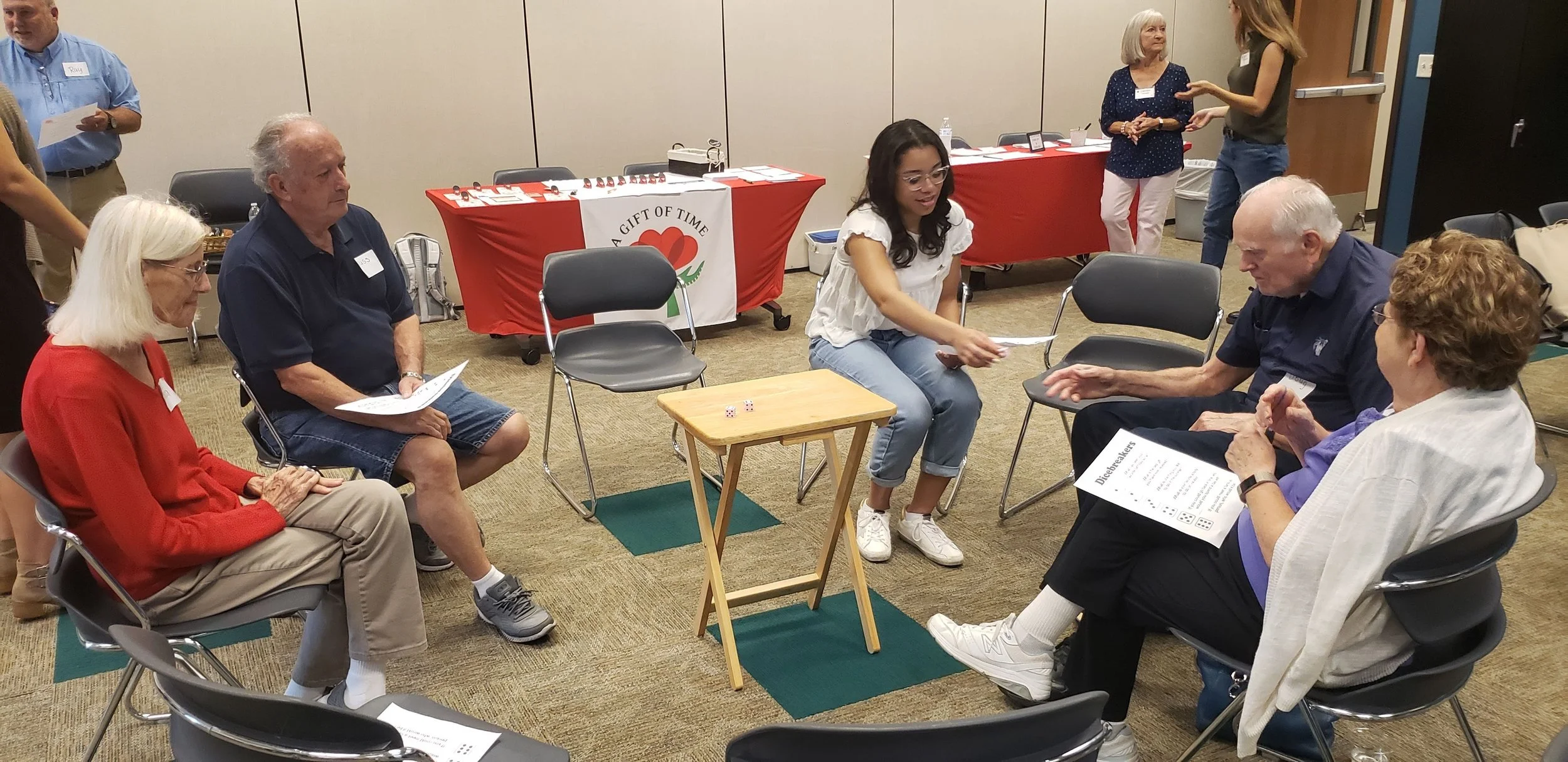 Group of elderly and young people participating in a bingo game, sitting in a circle with a small table with dice in the middle, in a room with informational tables and banners.