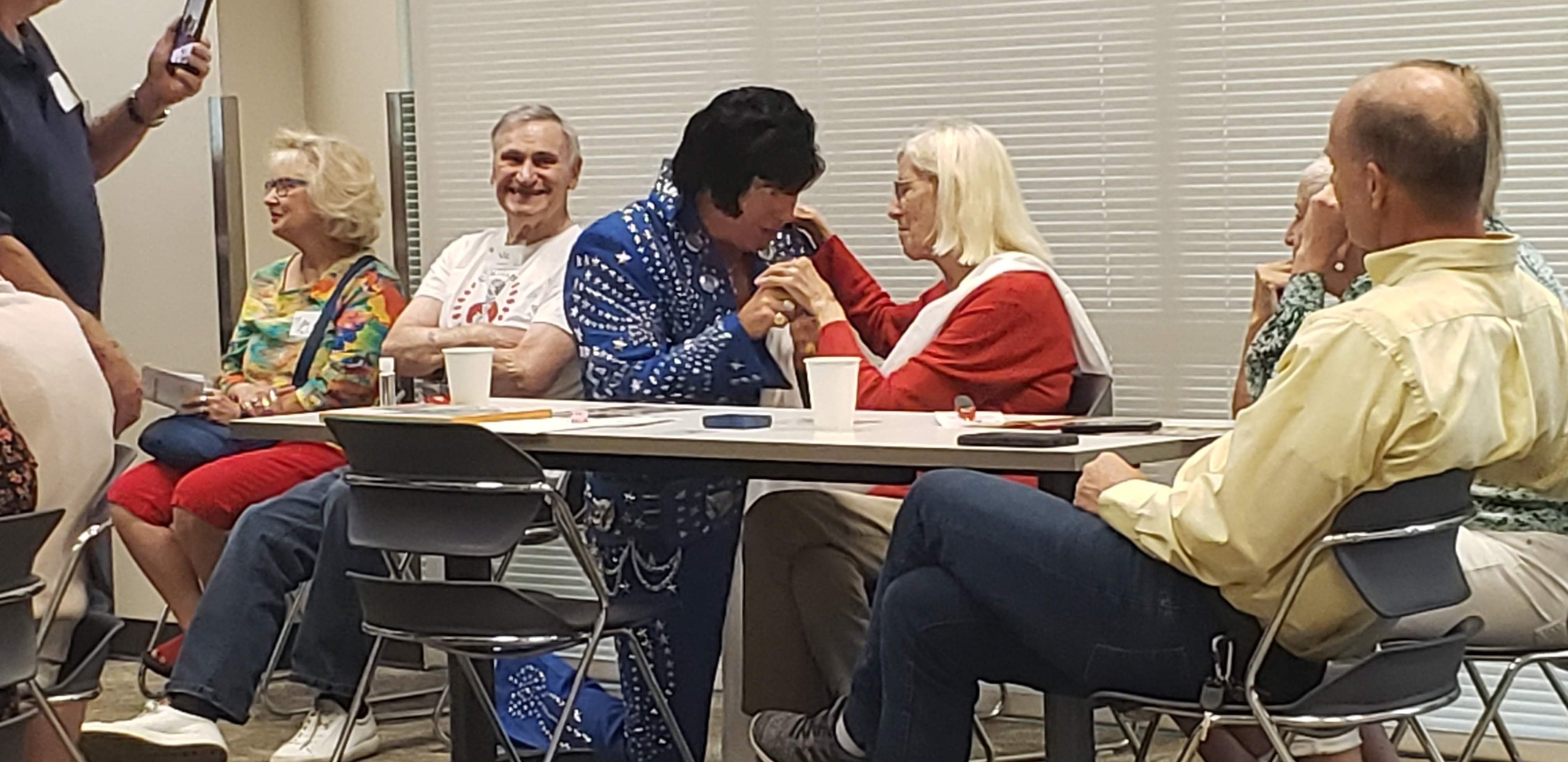 People sitting around a table at a meeting or gathering, some talking, some listening, with drinks and papers on the table.