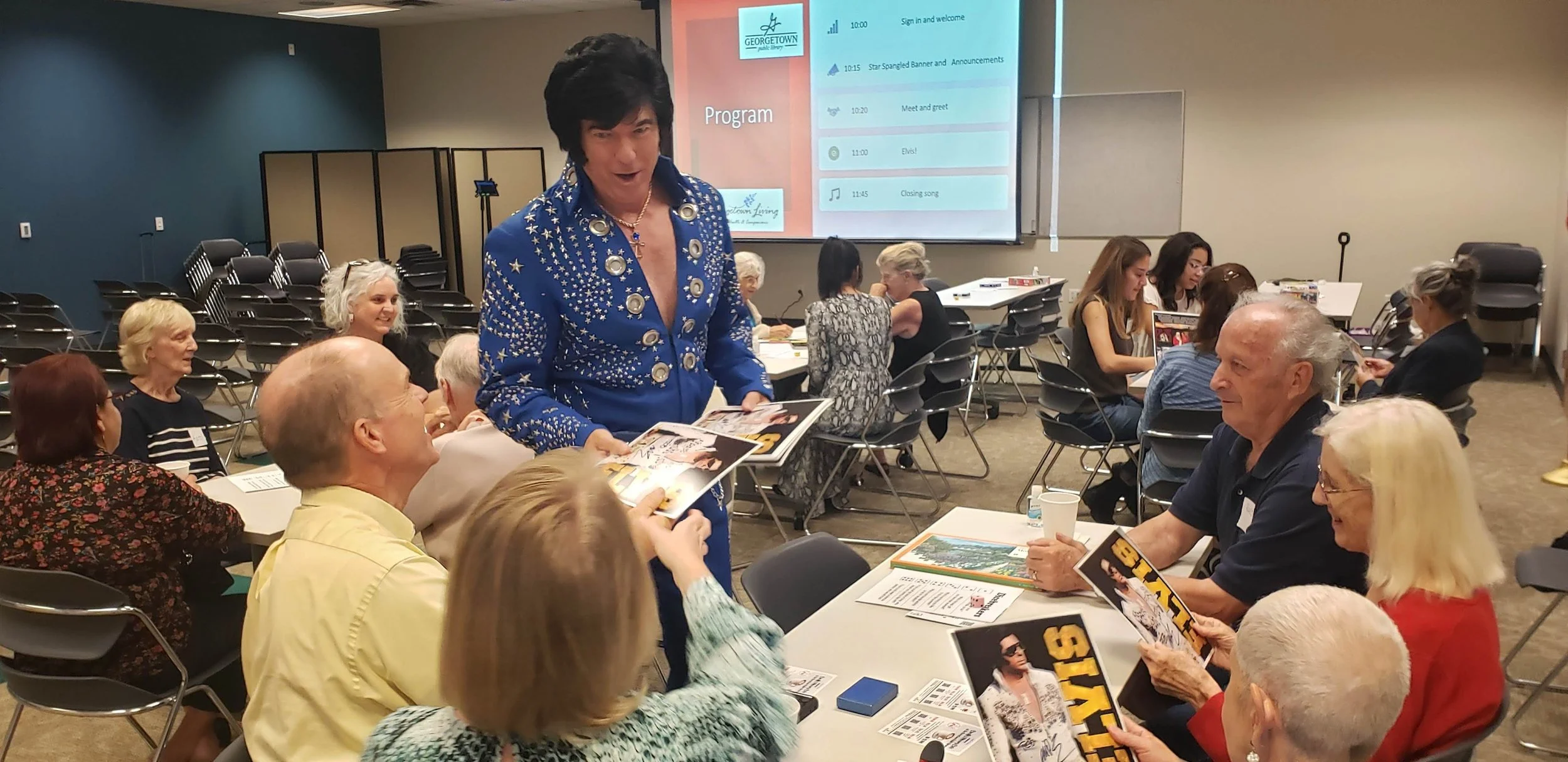 A group of people at a gathering in a conference room, with a person dressed as Elvis Presley handing out magazines and chatting with attendees.
