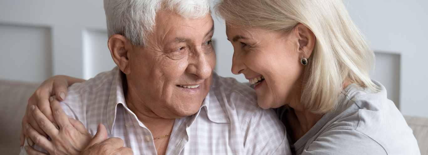 An elderly couple smiling and looking at each other closely in a cozy indoor setting.
