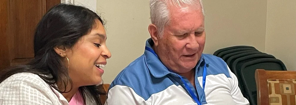 A woman and an older man sitting closely together, looking at something in front of them; she is smiling and he appears focused.