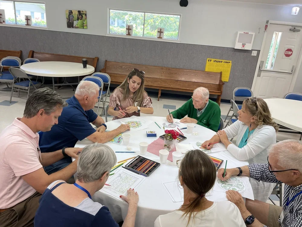 Group of adults sitting around a table, engaging in coloring activities with markers and papers in a community room.