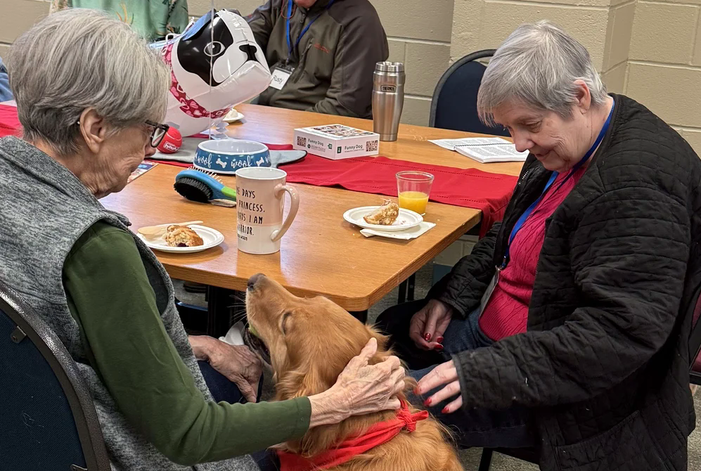 Two elderly women sitting at a table with a golden retriever dog, petting it and looking at each other, during a gathering with food and drinks.