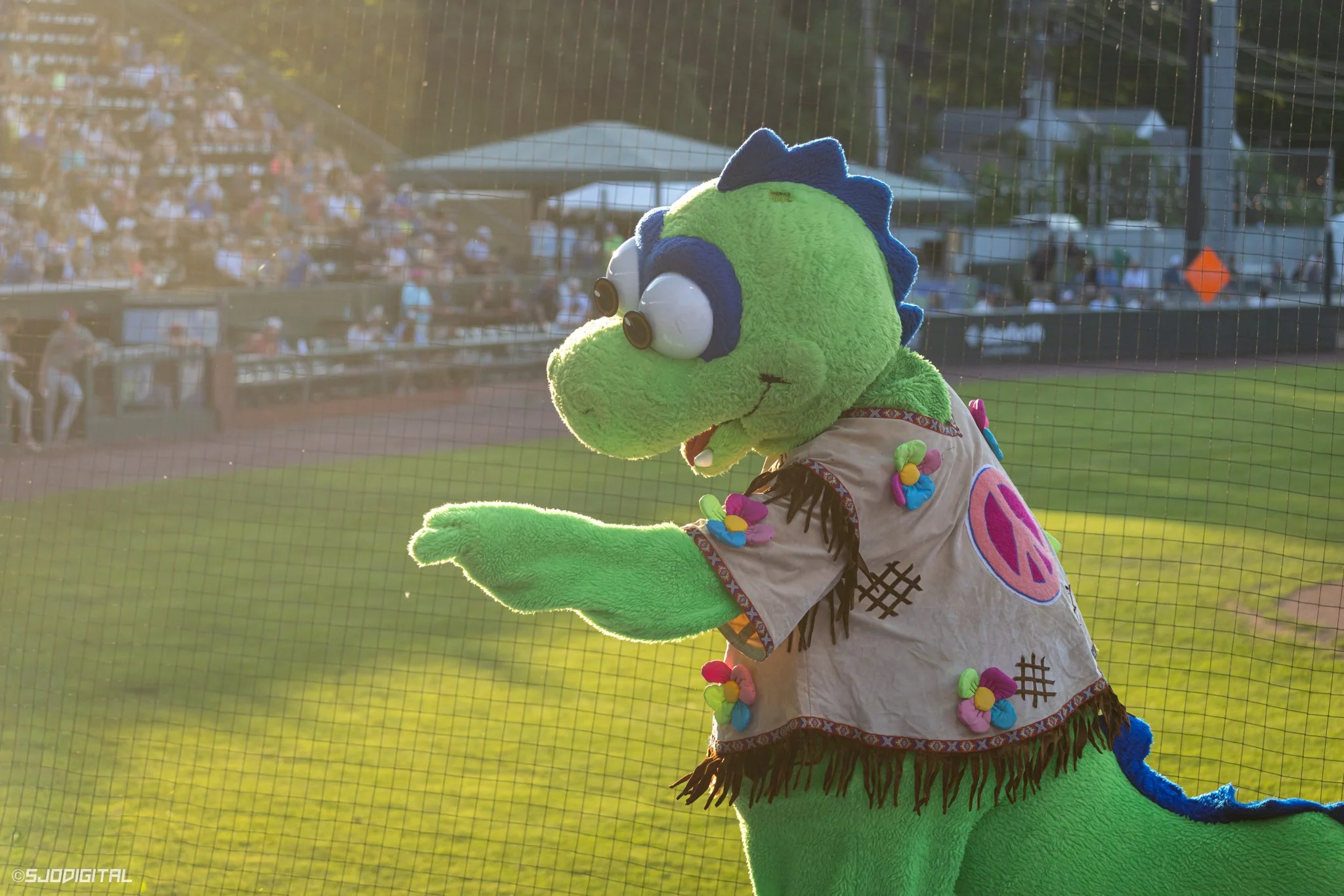 A person dressed in a bright green dinosaur costume at a sporting event, standing near a fence on the field with a crowd visible in the background.