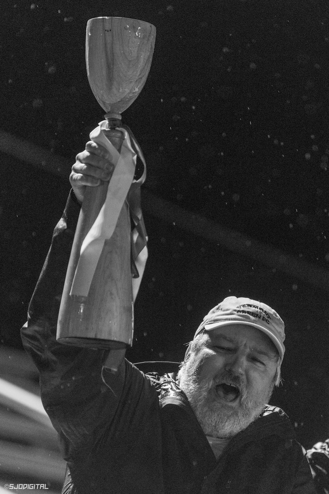 An older man with a beard, wearing a cap and jacket, cheering and raising a trophy in the air with an expression of joy.