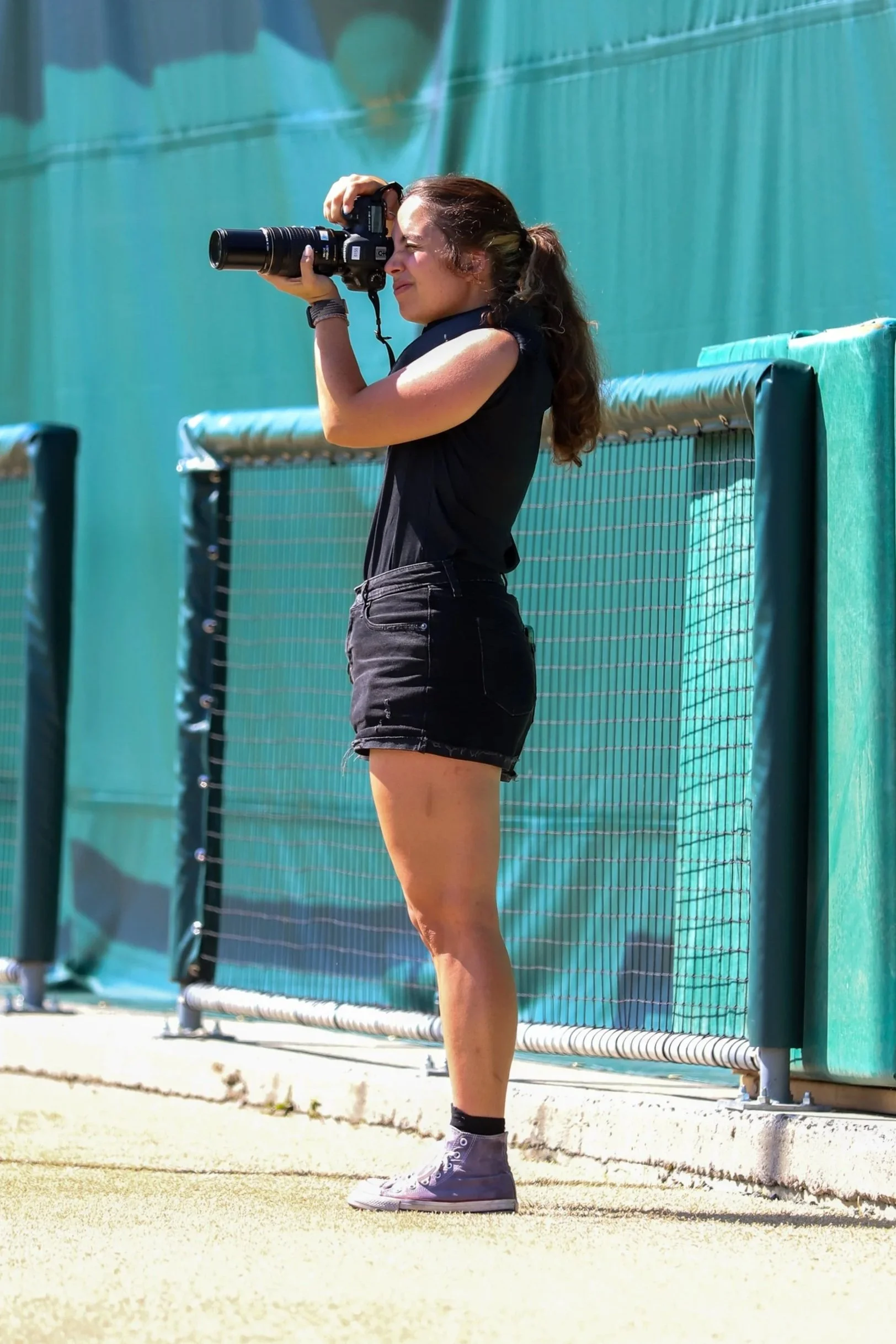 A woman standing on a tennis court, taking a photo with a camera, wearing a black sleeveless top, black shorts, and gray sneakers, next to green fencing.