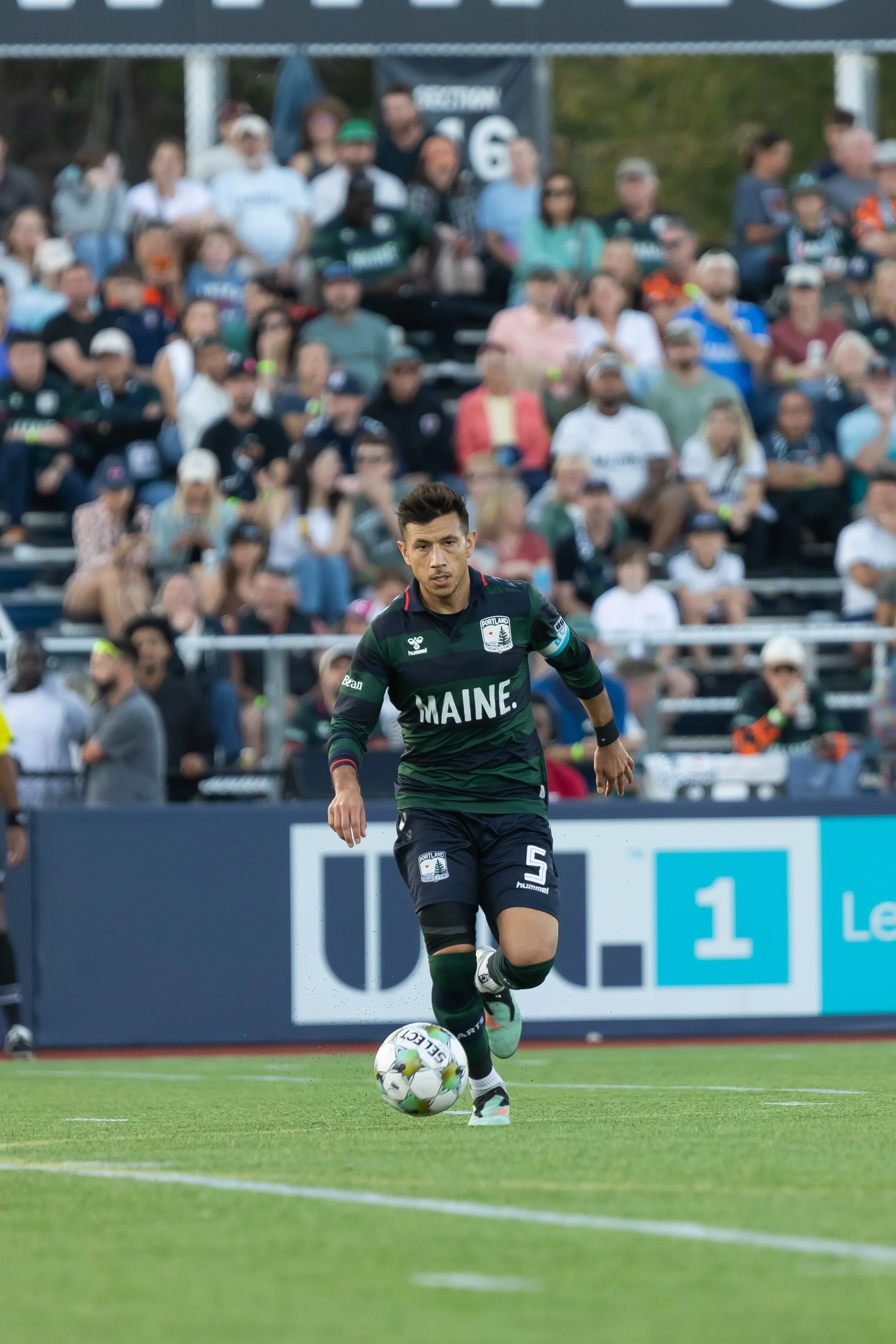A soccer player in a black and green uniform is running with a ball on a soccer field. The player is in focus, with a crowd of spectators blurred in the background.