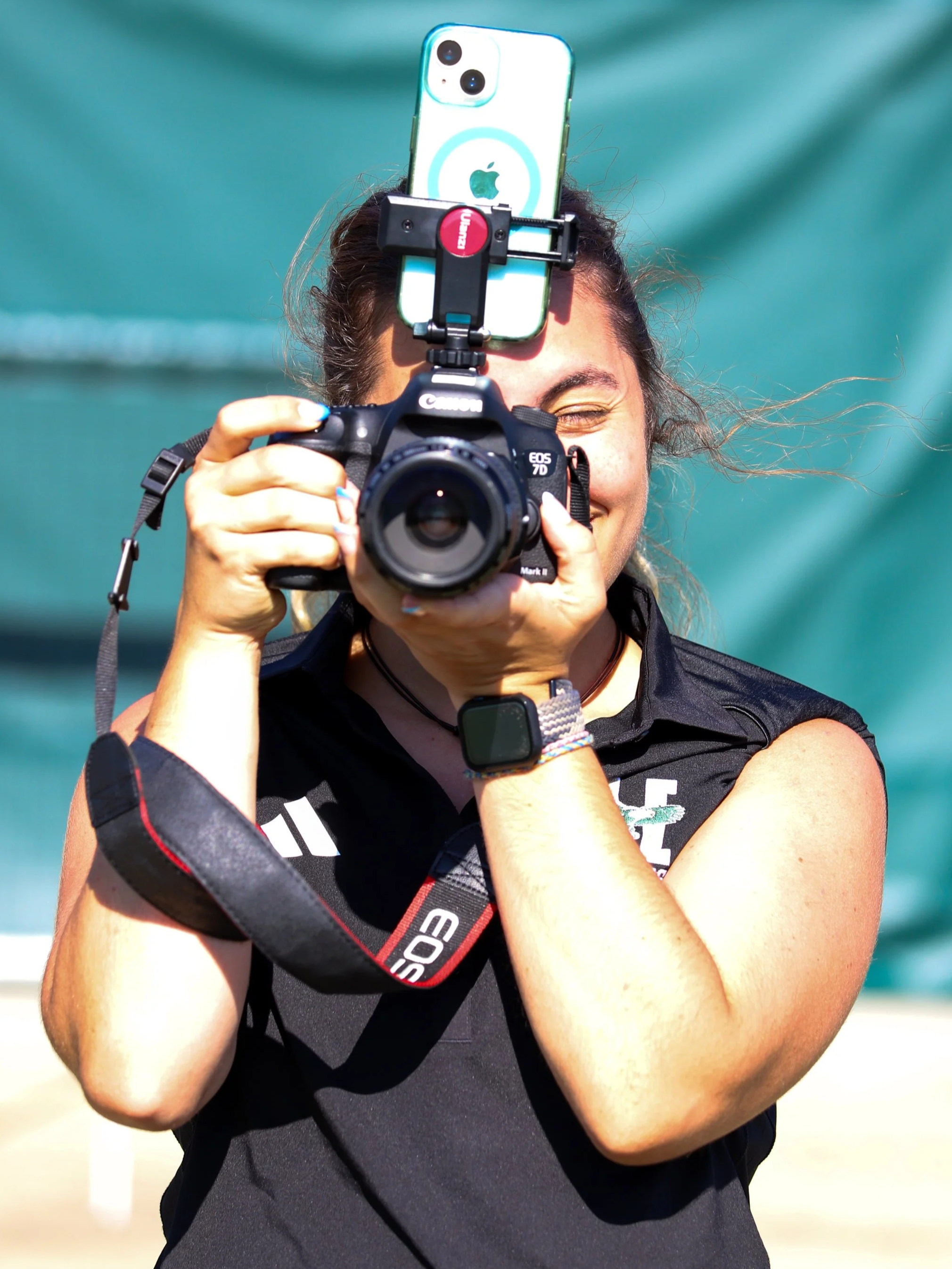 A woman taking a selfie with a camera mount attached to her face, with a smartphone on top, in an outdoor setting with a teal background.