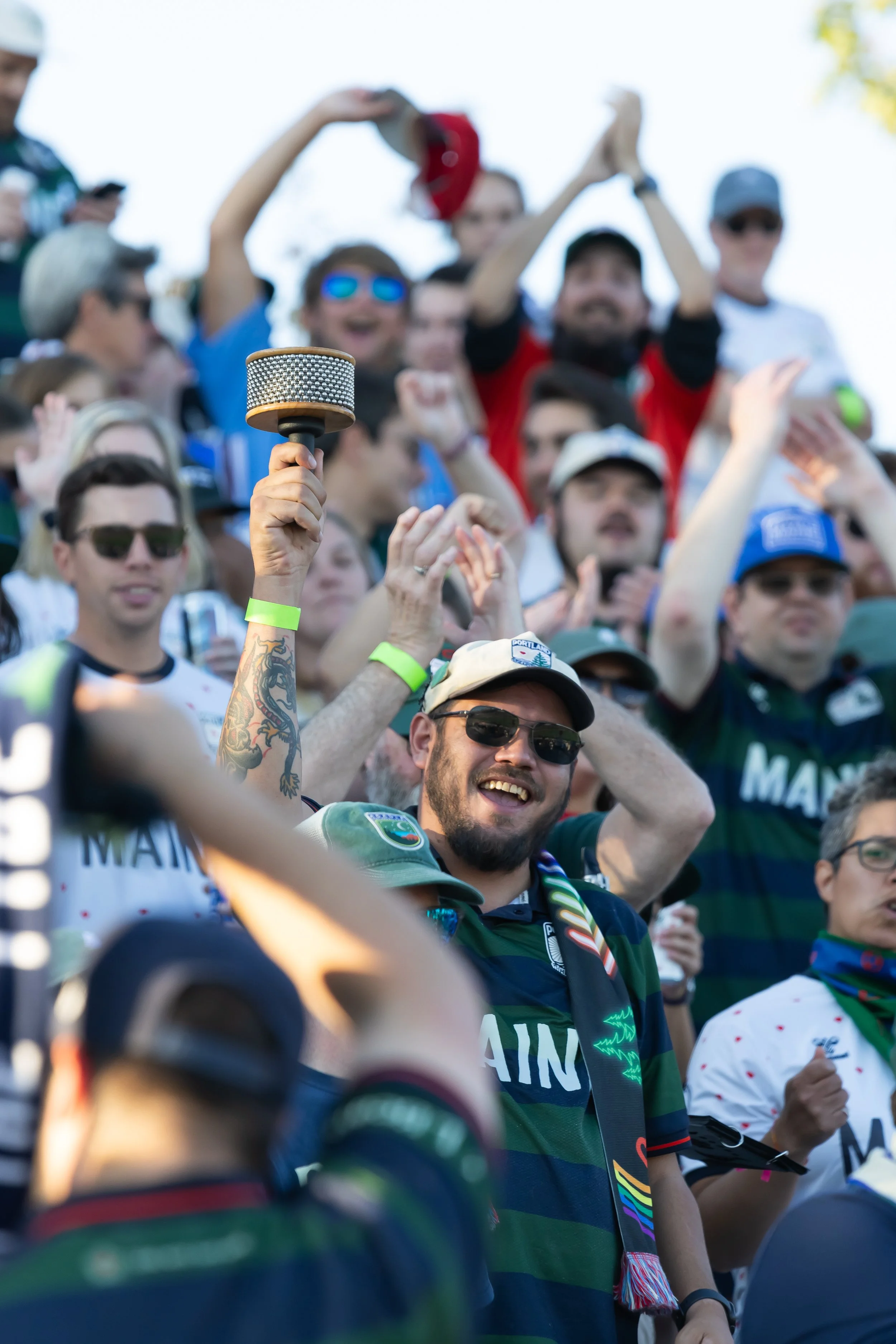 A crowd of cheering sports fans, many wearing sunglasses and team jerseys, holds a microphone and celebrates during a game outdoors on a sunny day.