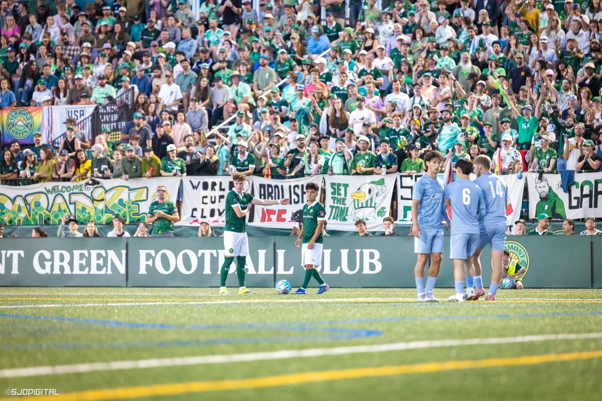 A soccer match with players on the field and a crowd of spectators in the stands, some holding flags and banners supporting their team.