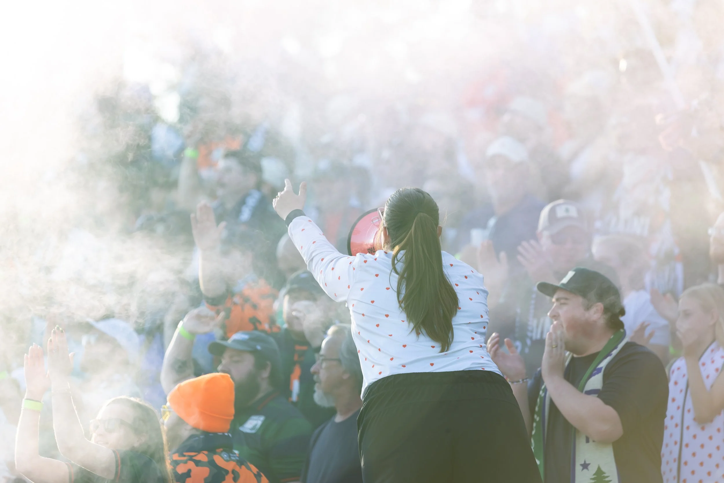 A crowd of people celebrating outdoors with some raising their hands, and a girl with long hair tied back wearing a white shirt with red hearts, standing in the center.