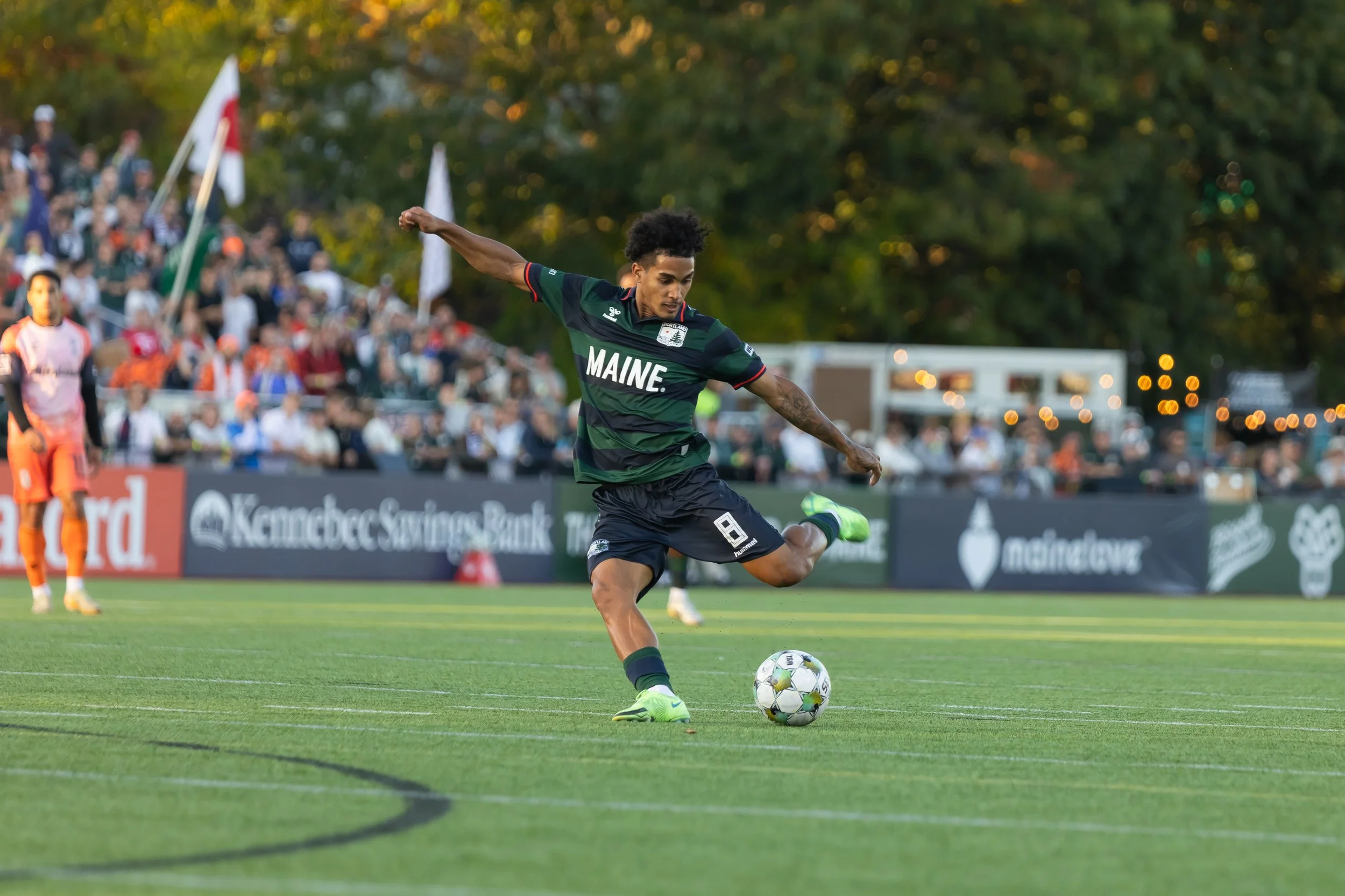 A soccer player wearing a Maine team jersey with the number 8 kicks a ball during a match on a grass field, with a crowd of spectators and trees in the background.
