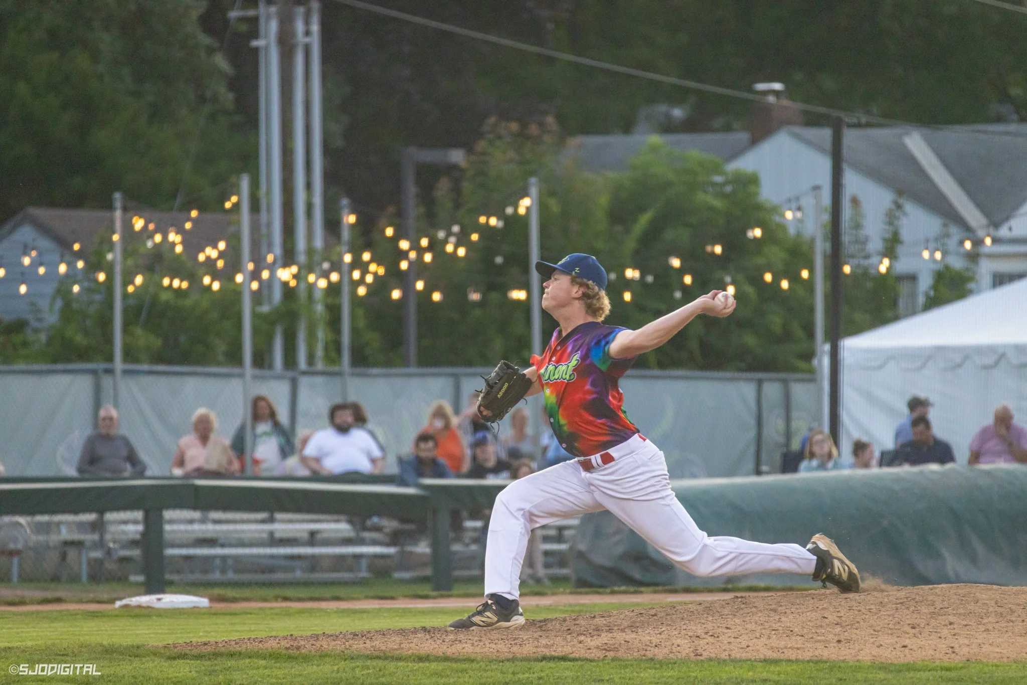 A baseball player in a colorful jersey and white pants is pitching on a baseball field during evening with spectators watching behind a fence, and string lights and houses in the background.