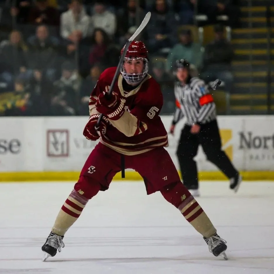 Ice hockey player in maroon and gold uniform and helmet, holding a stick, on the ice during a game, with a referee and spectators in the background.