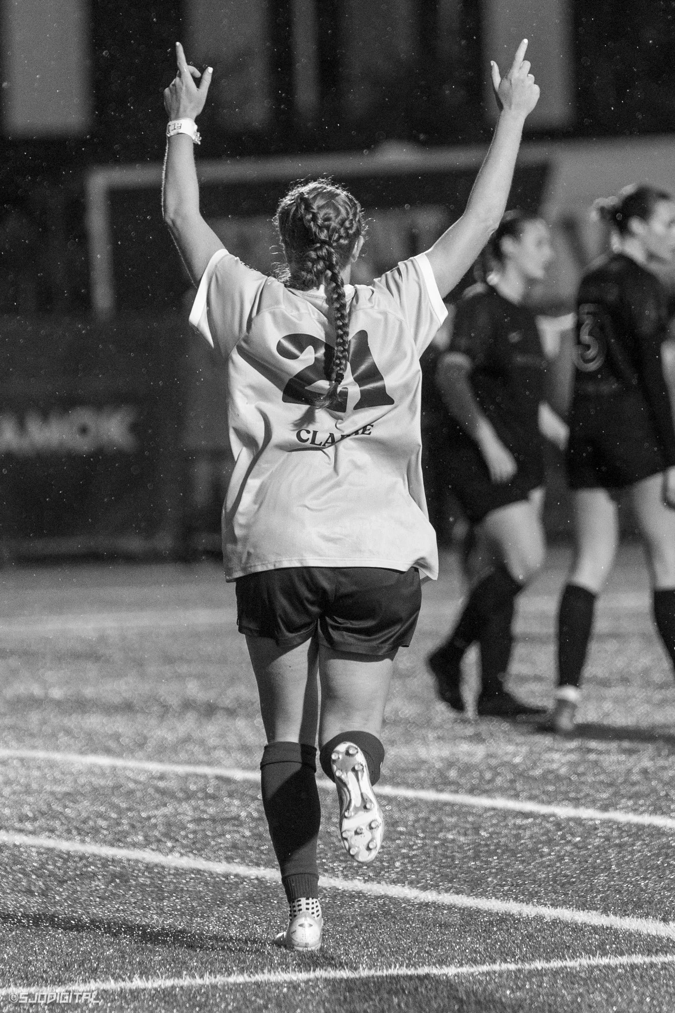 A female soccer player wearing a jersey with the number 21 and the name Claire, running on a wet field with arms raised, while other players are in the background.