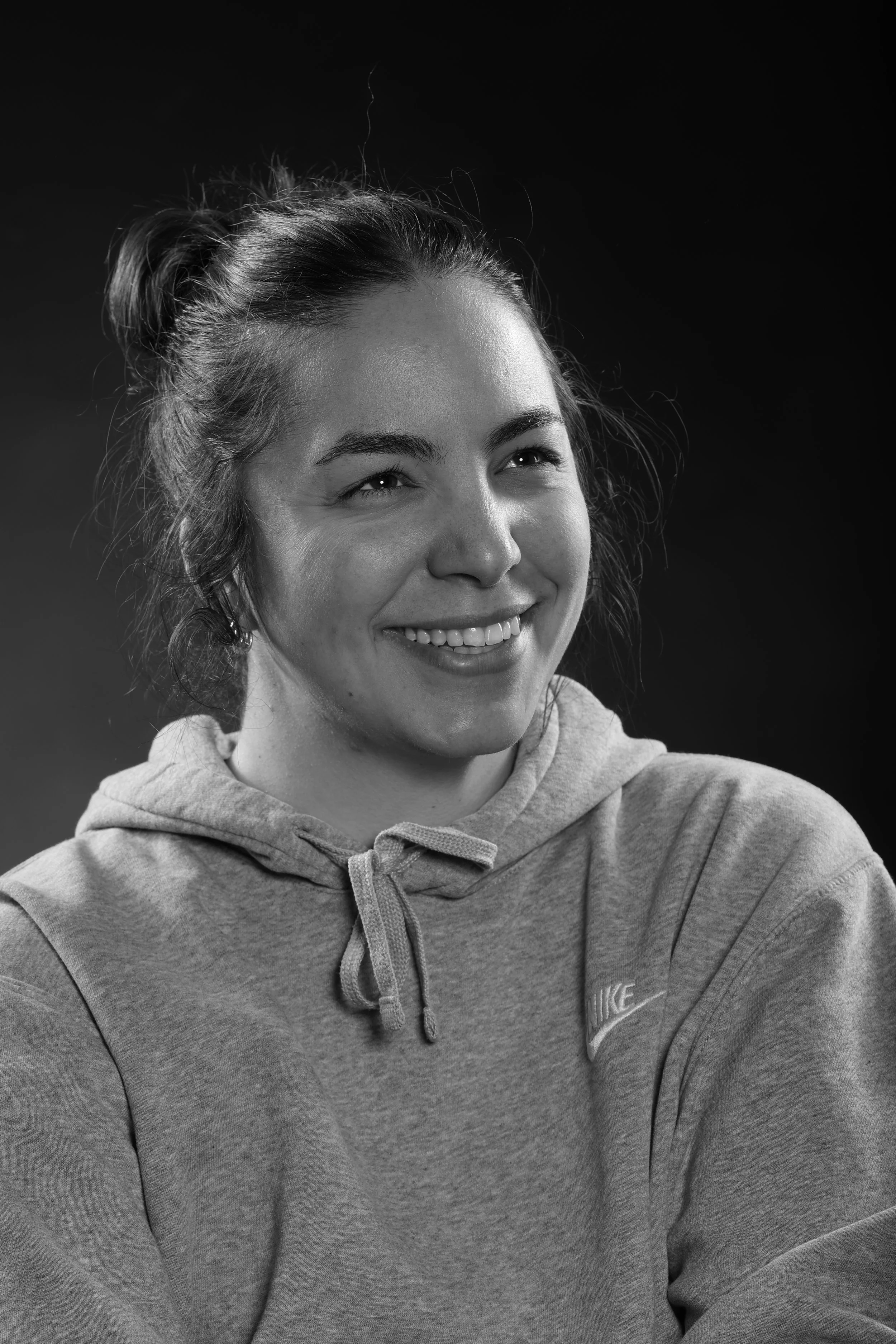 A black and white photo of a young woman smiling, wearing a hoodie, with her hair in a bun.