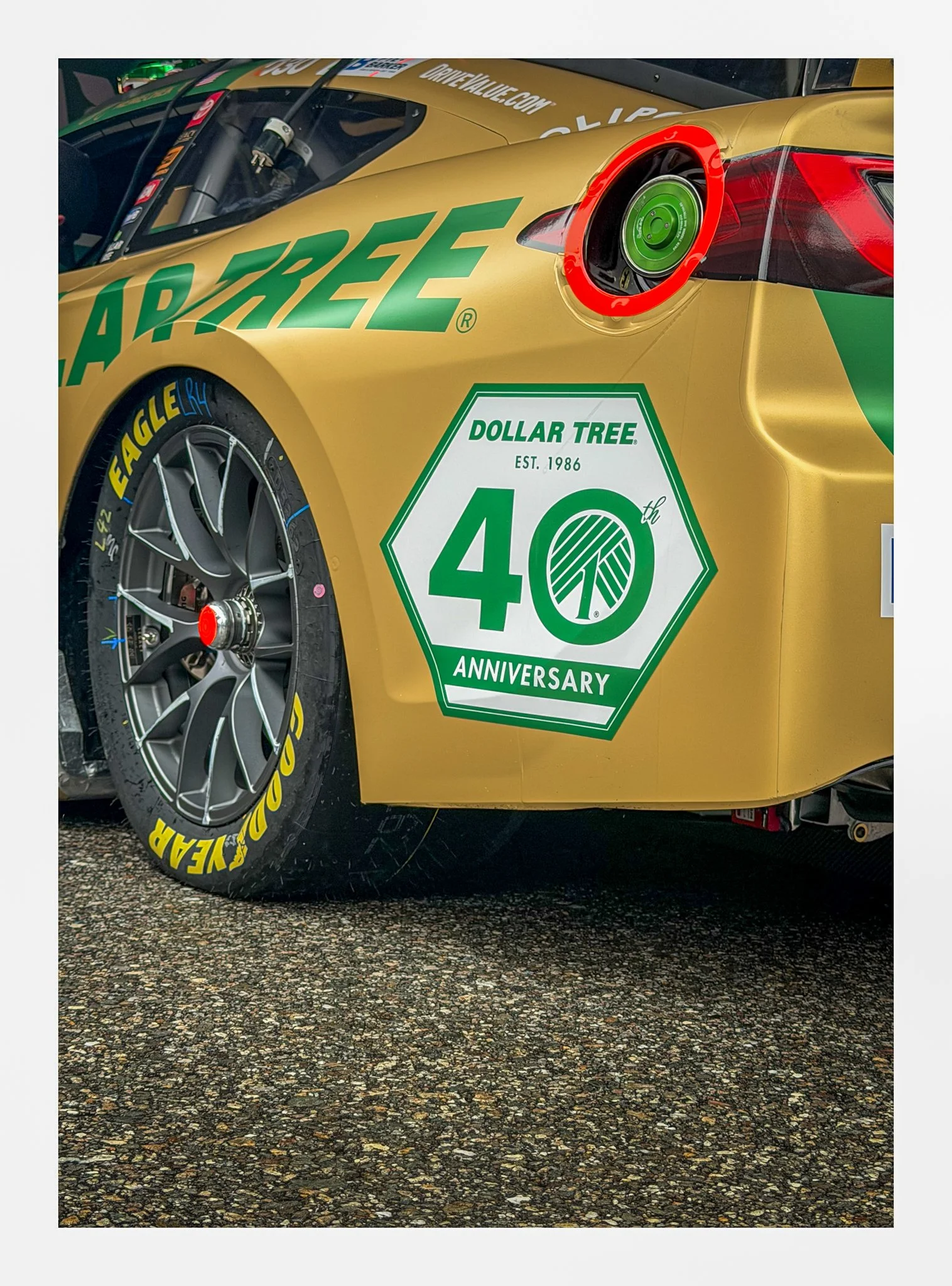 Close-up of a yellow race car with green and red sponsor logos, featuring a yellow and black tire with 'Goodyear' written in yellow text, and a green fuel cap surrounded by a red ring. The car has a large white and green decal celebrating the 40th an