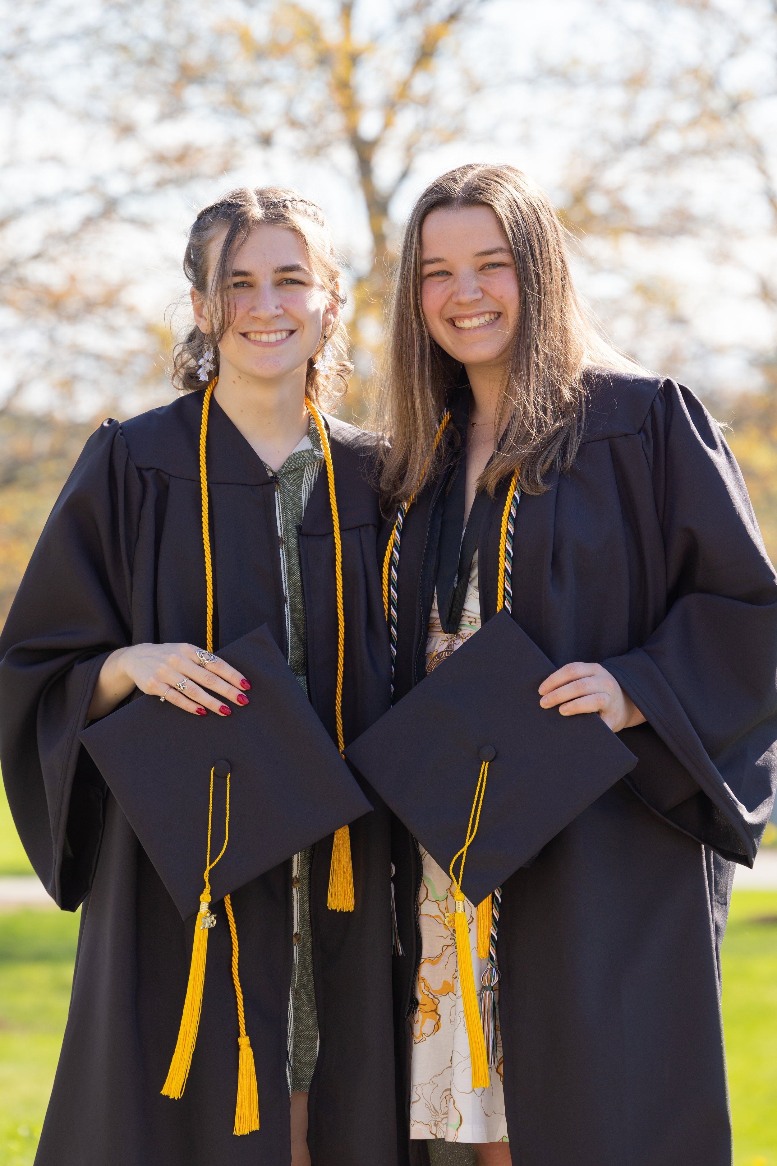 Two young women in graduation caps and gowns pose outdoors with trees in the background.