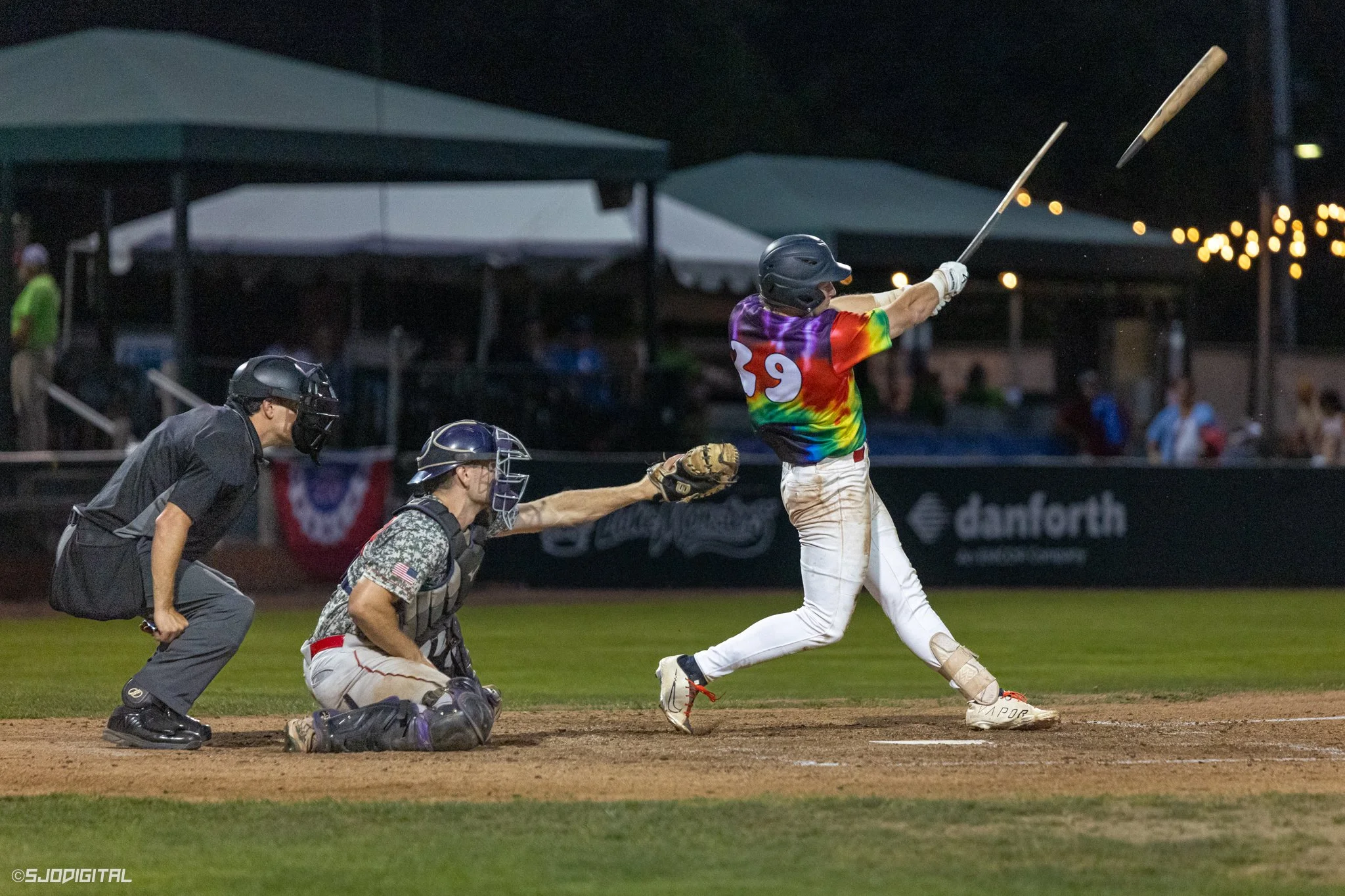 A baseball game at night, showing a player in a rainbow-colored jersey hitting the ball, with an umpire and catcher behind him, and the ball flying through the air.