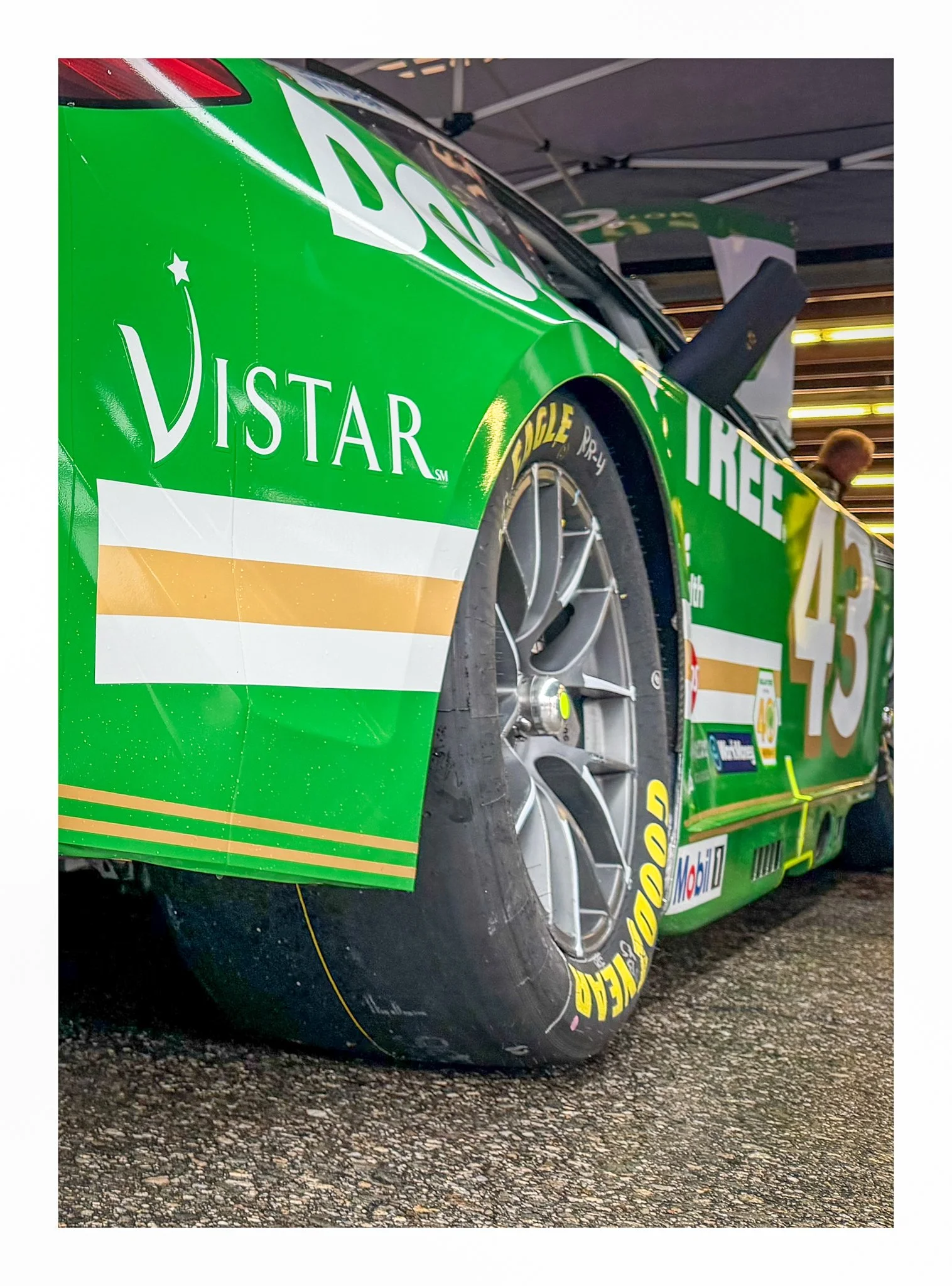 Close-up of a green stock car at a racing event, showing the front side with visible tire and part of the hood, under a tent with someone in the background.