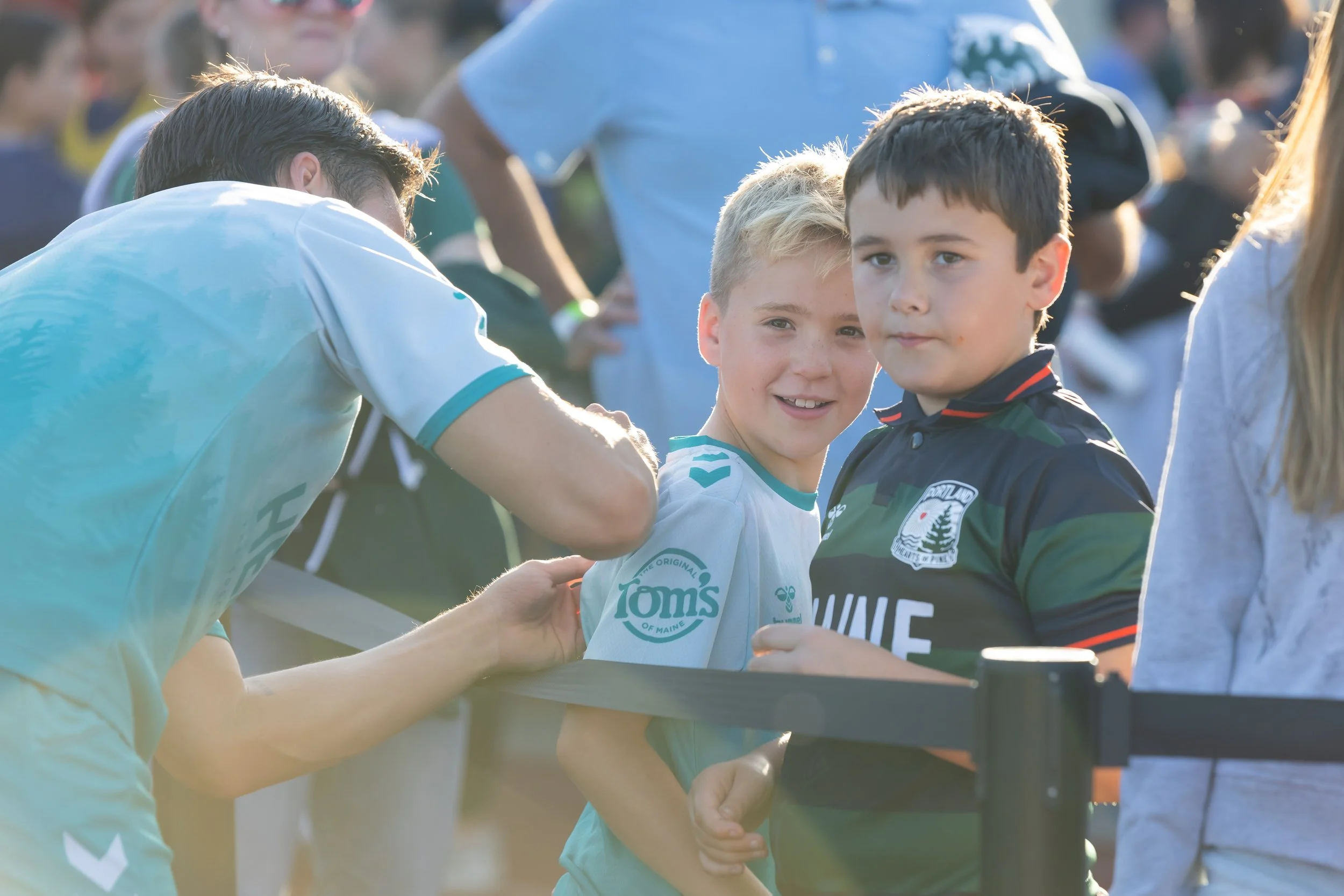 Two young boys in sports jerseys shaking hands with a man during an outdoor sports event, with other people and players in the background.