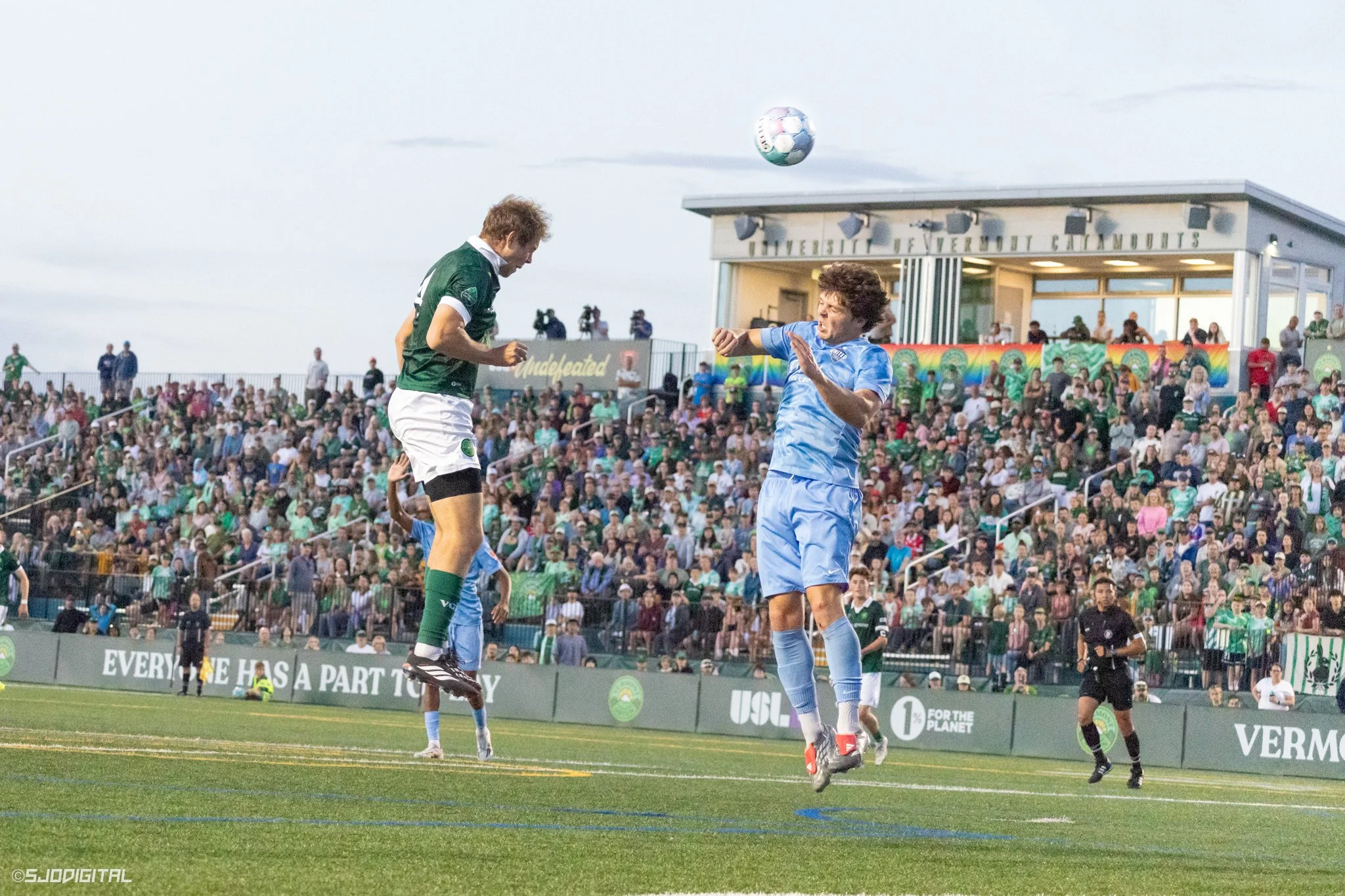 Soccer players in action during a match, with one player jumping to head the ball while another attempts to block. Spectators watch from the stands in the background.