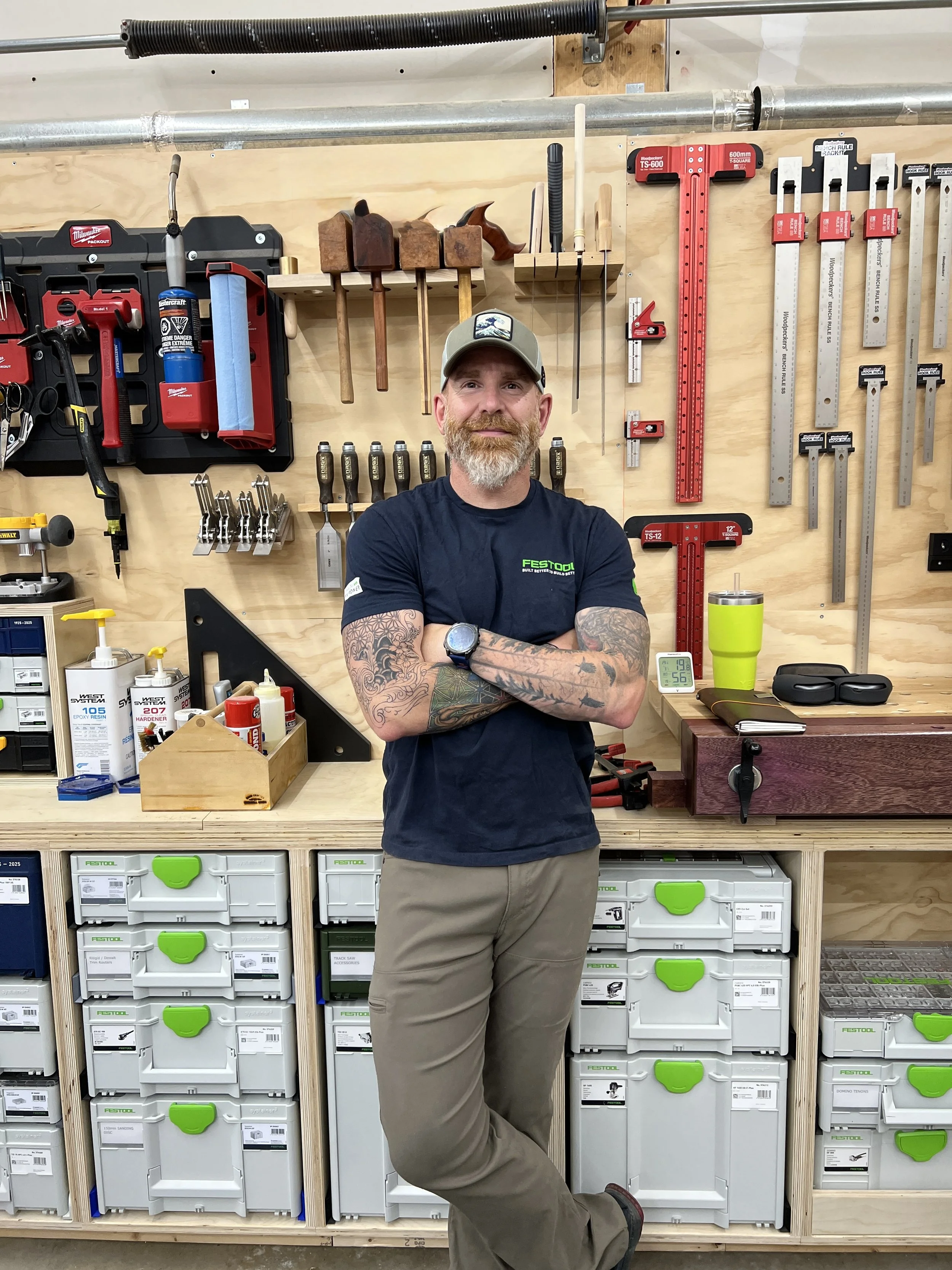 A man with a beard and tattoos, wearing a baseball cap and a dark blue T-shirt with 'Festool' written on it, stands in a woodworking workshop with arms crossed and one leg rested on a lower surface. Behind him are various woodworking tools hanging on a wooden wall, including hammers, saws, rulers, and clamps. There are storage drawers with green handles on a workbench.
