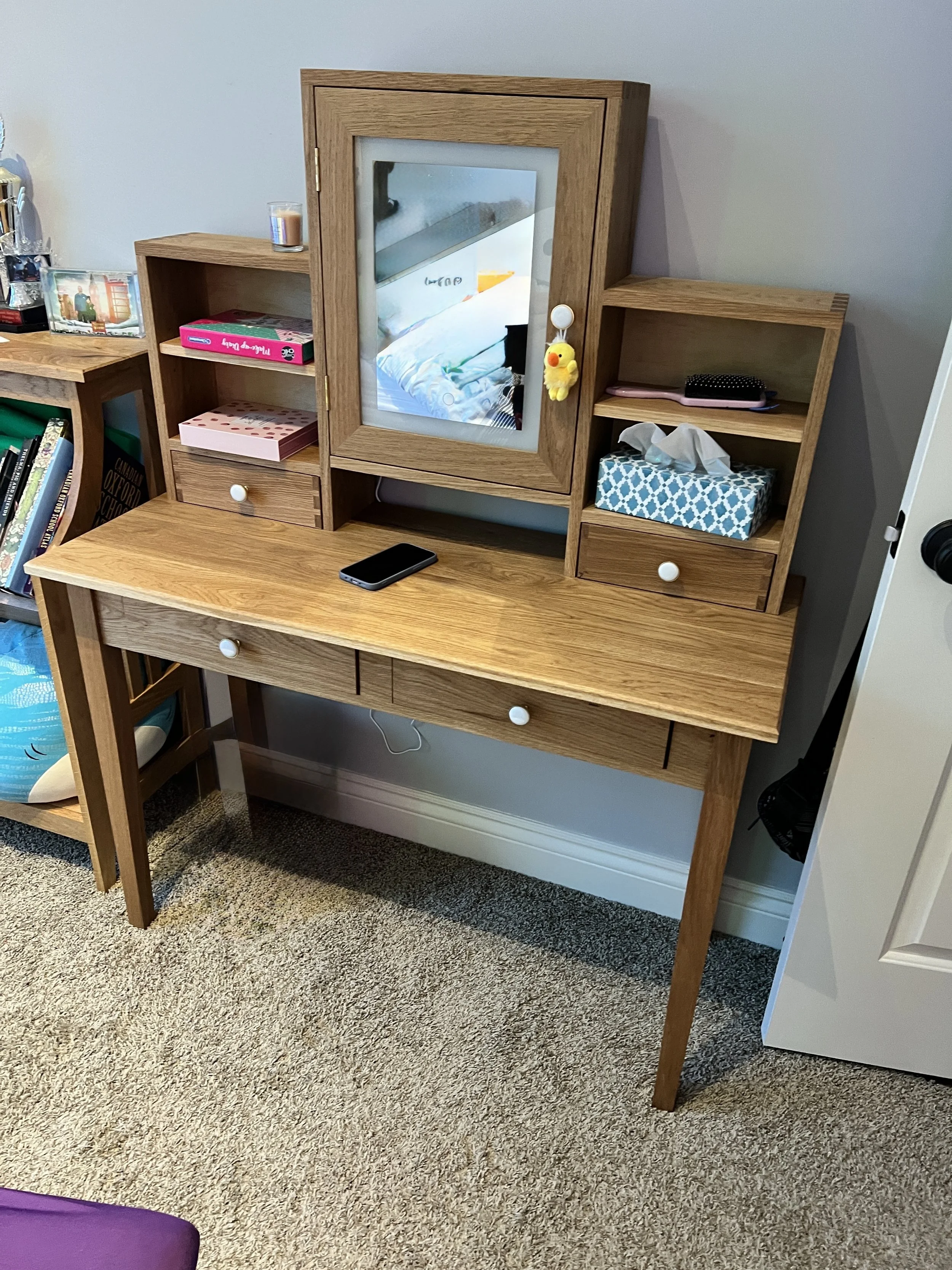 Wooden vanity with a mirror, three open shelves, and two drawers, placed against a wall on a carpeted floor. Items on the shelves include a pink box, tissues, and a black hairbrush. A phone is on the top surface of the vanity. To the left, a bookshel
