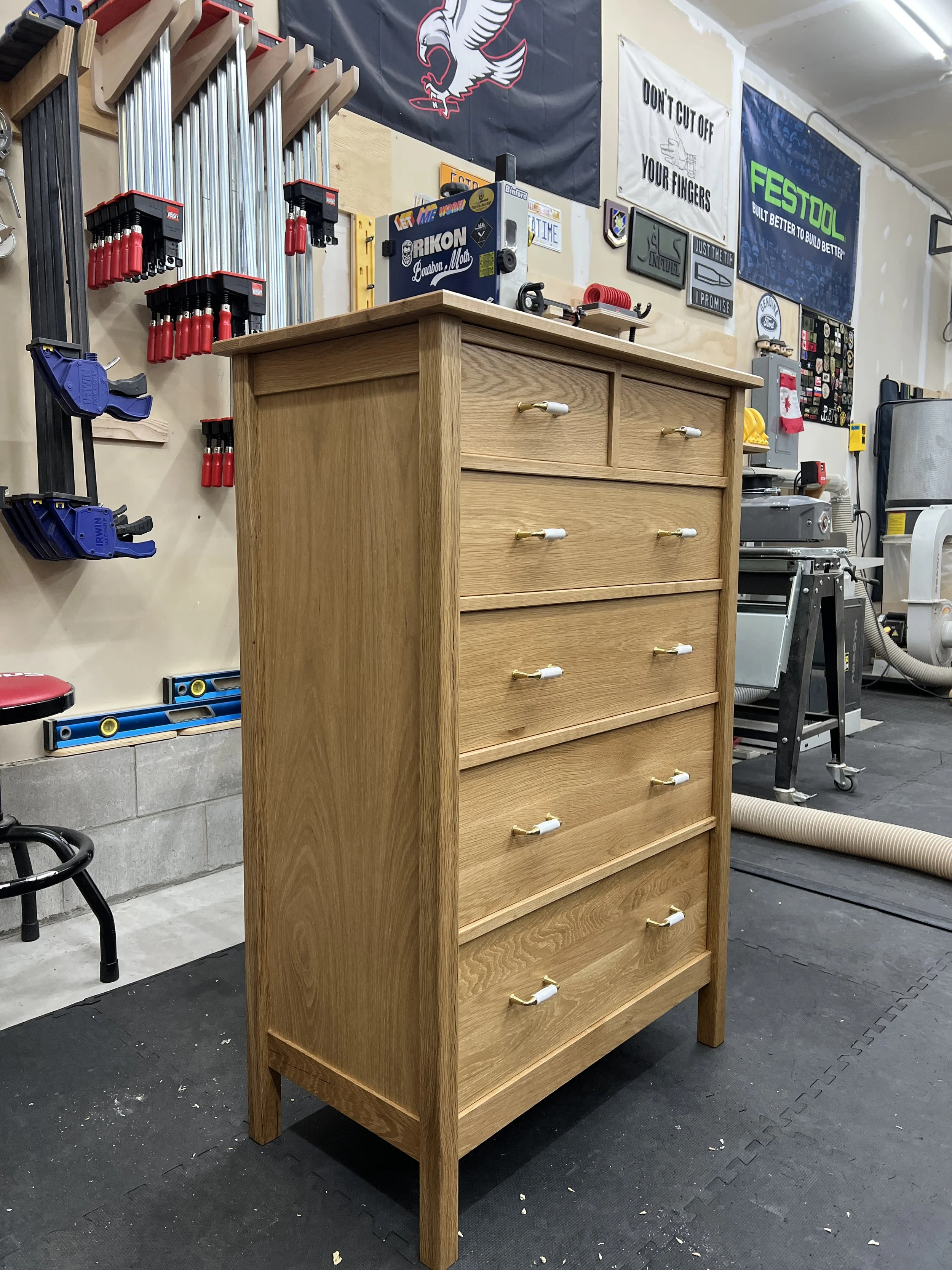 A tall wooden dresser with six drawers and white handles with gold accents in a workshop. The workshop has various tools, signs, and equipment on the walls and floor.
