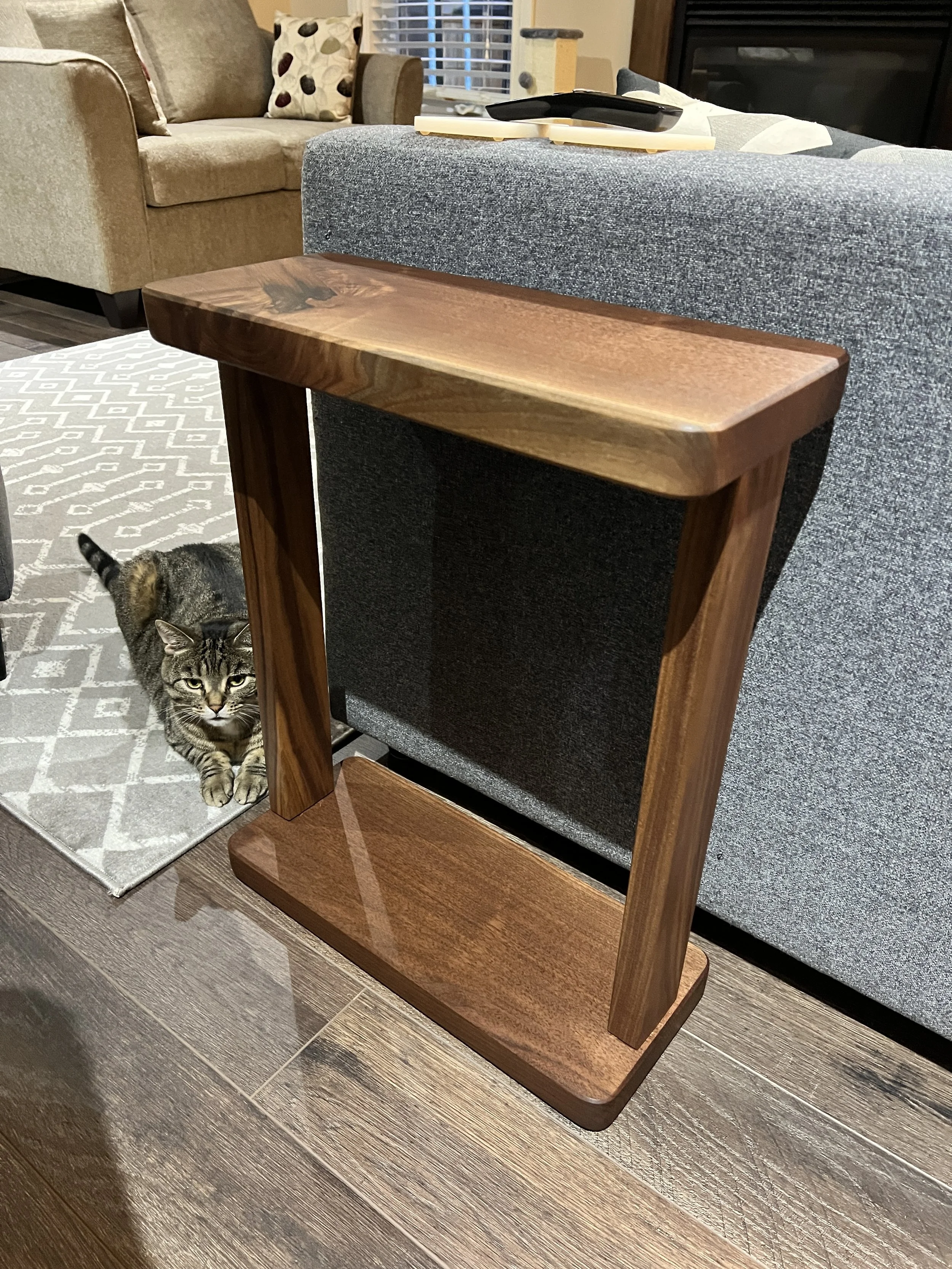 A wooden side table placed in front of a gray couch in a living room. A tabby cat with distinctive black, brown, and gray fur is lying on a gray and white patterned rug nearby, looking at the camera. In the background, there are cushions on the couch
