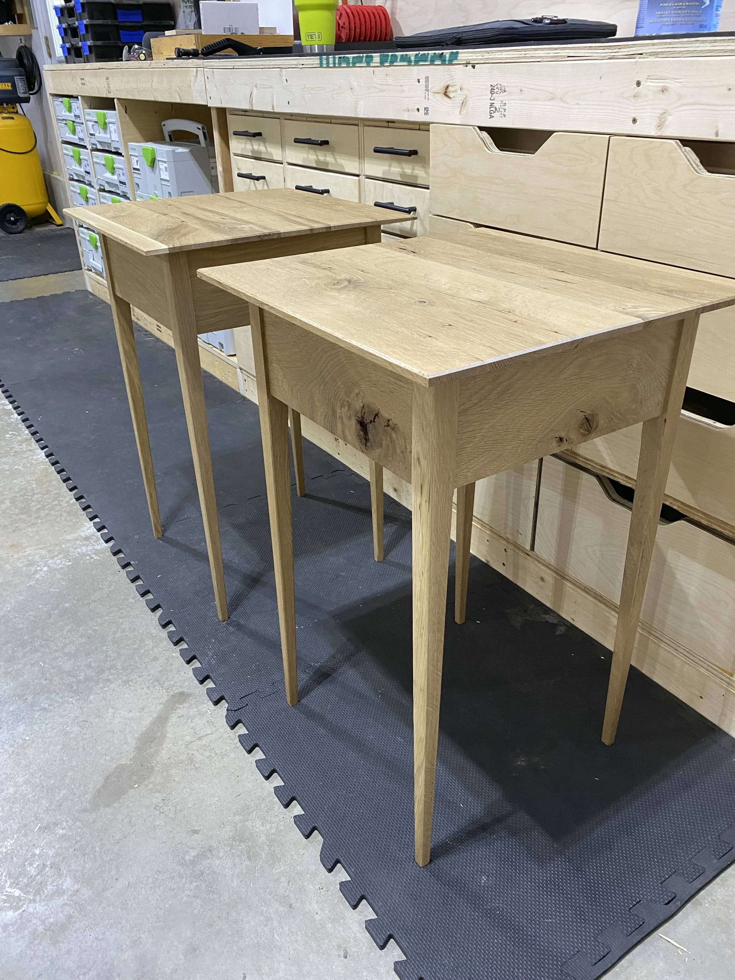 Two unfinished wooden tables with four legs each, placed in a workshop with storage drawers and tools in the background.