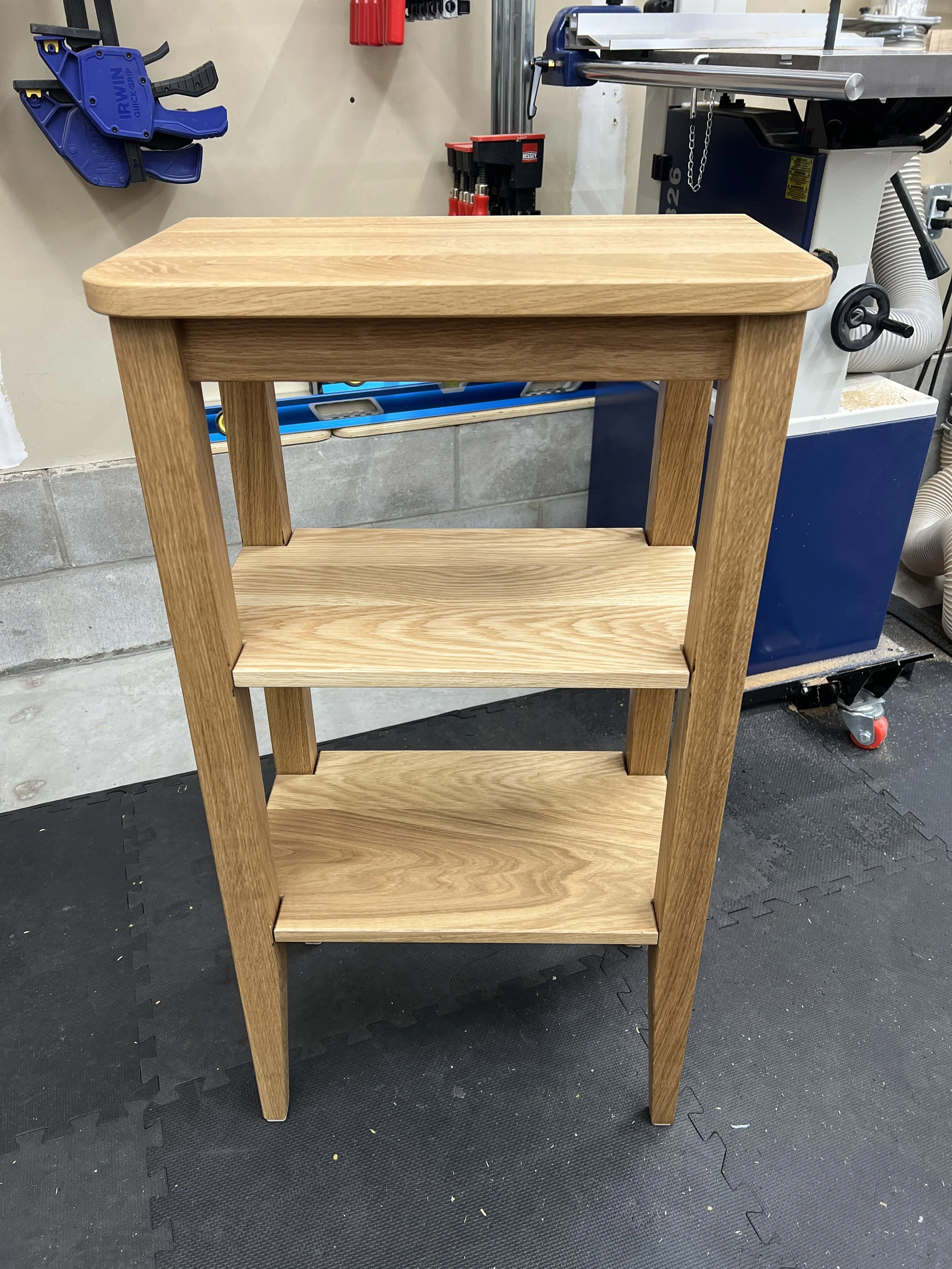 A wooden shelf unit with four shelves stands on a black rubber mat in a workshop. Various tools and equipment are visible in the background.