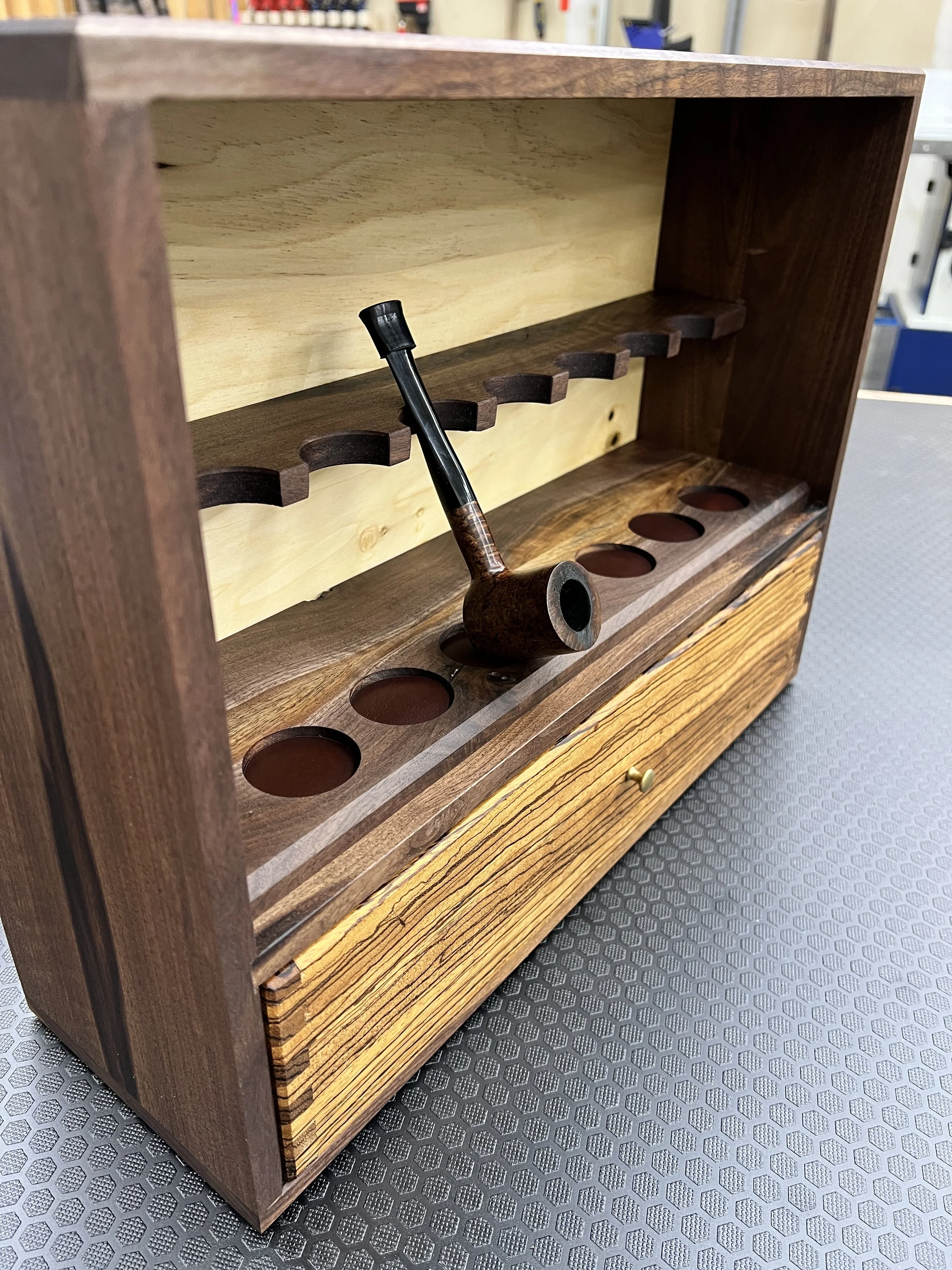 A wooden bookshelf with a pipe resting on its middle shelf, and round holes in the bottom shelf, on a black textured surface.